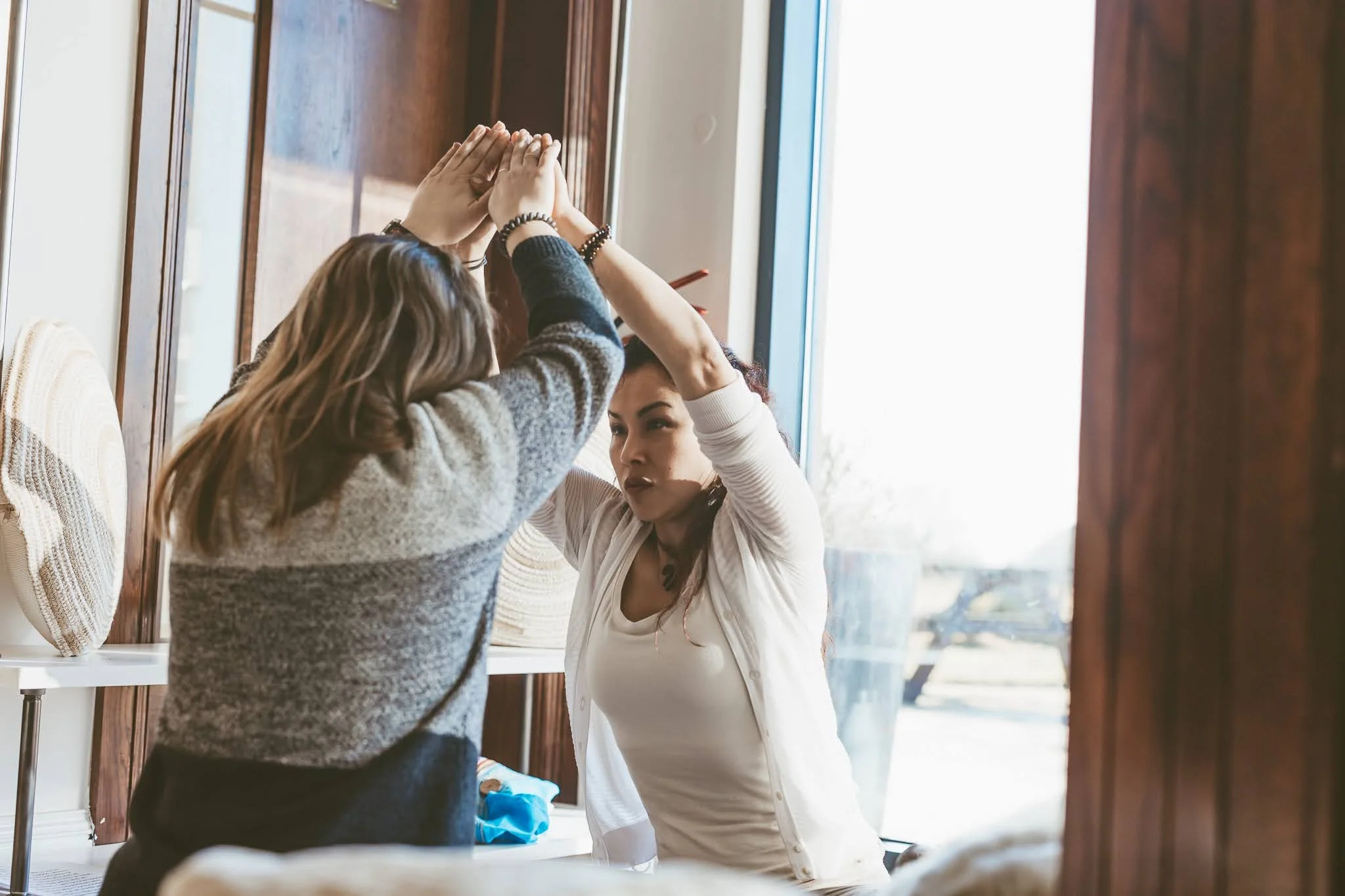 Two women are practicing self-defense techniques in a room with wooden walls and a large window, one woman is blocking the other's arm as part of a training exercise.