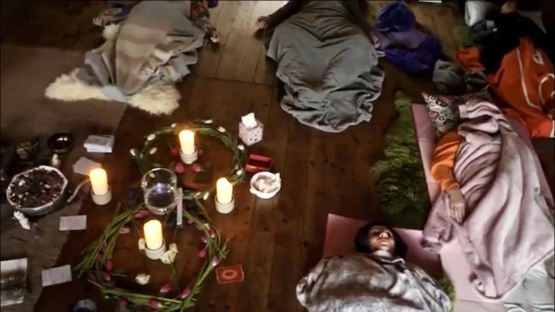 People sleeping on the floor next to lit candles, blankets, and scattered belongings during what appears to be a memorial or vigil.