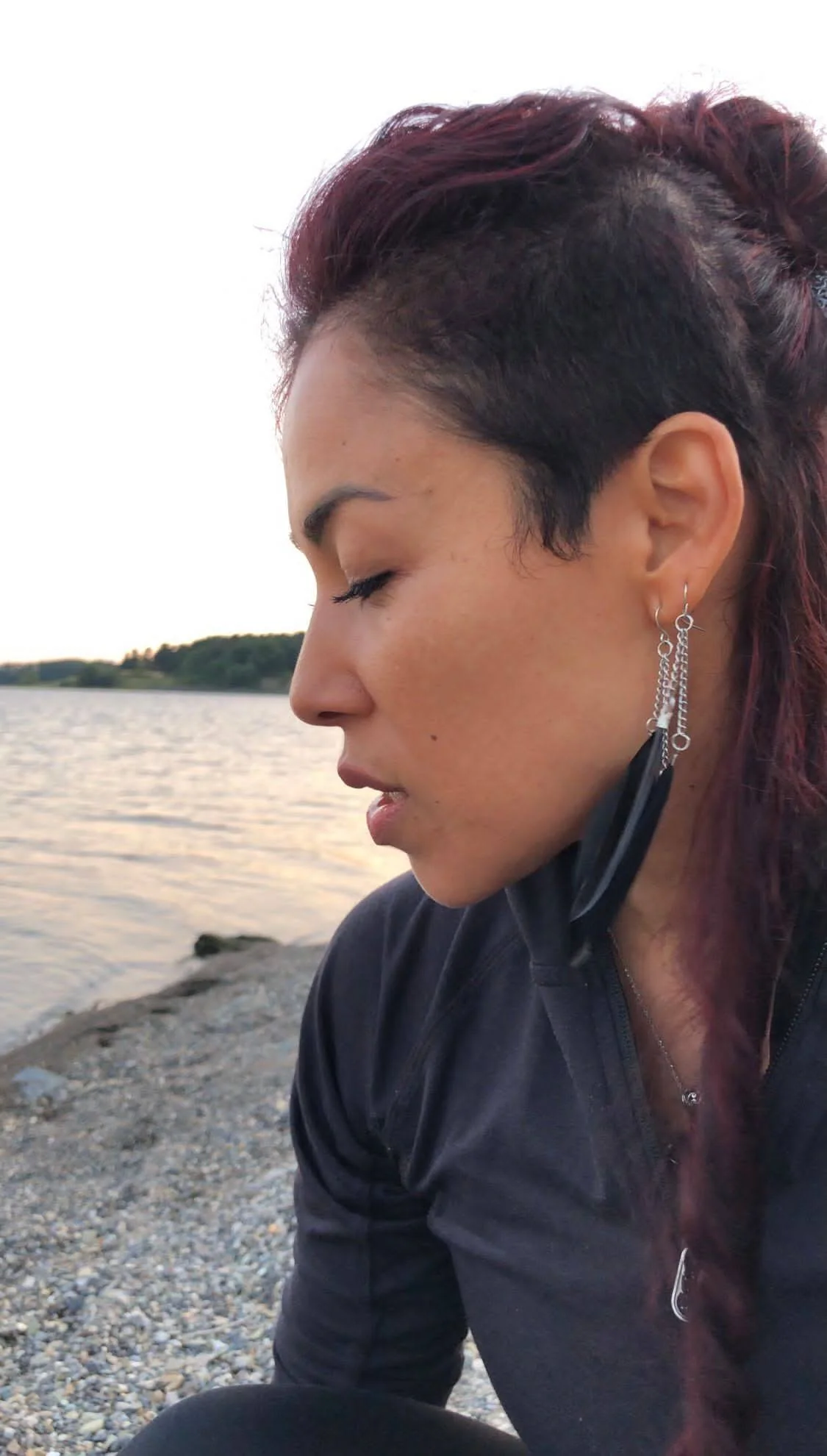 Side profile of a woman with short, dark, and reddish hair, wearing earrings and a black jacket, sitting near a body of water with a distant shoreline, during sunset.