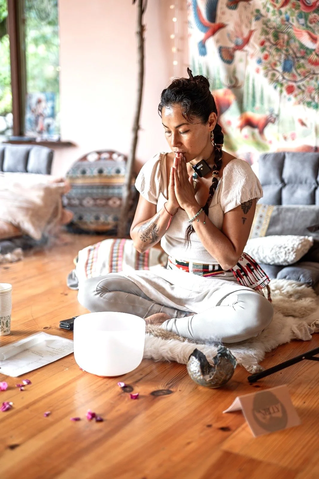 A woman practicing meditation or prayer in a cozy room with colorful decorations, including a tapestry with animal designs, a large window, and scattered rose petals on the wooden floor.