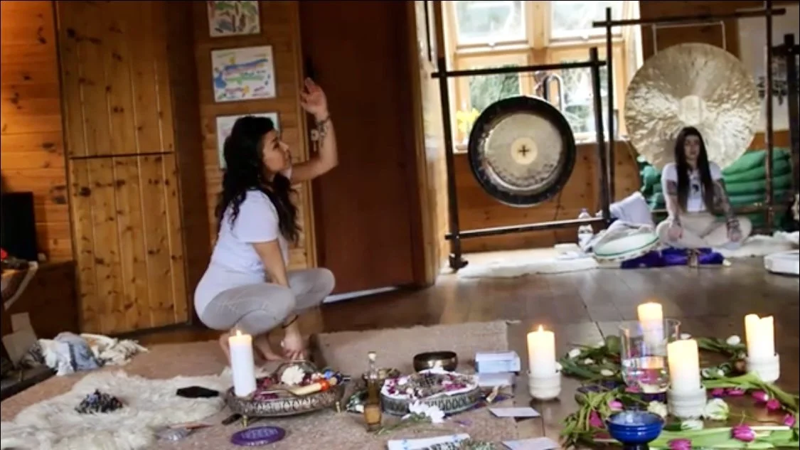 Two women sitting on the floor with candles, meditation supplies, and decorations in a cozy wooden room, one practicing a meditation pose.