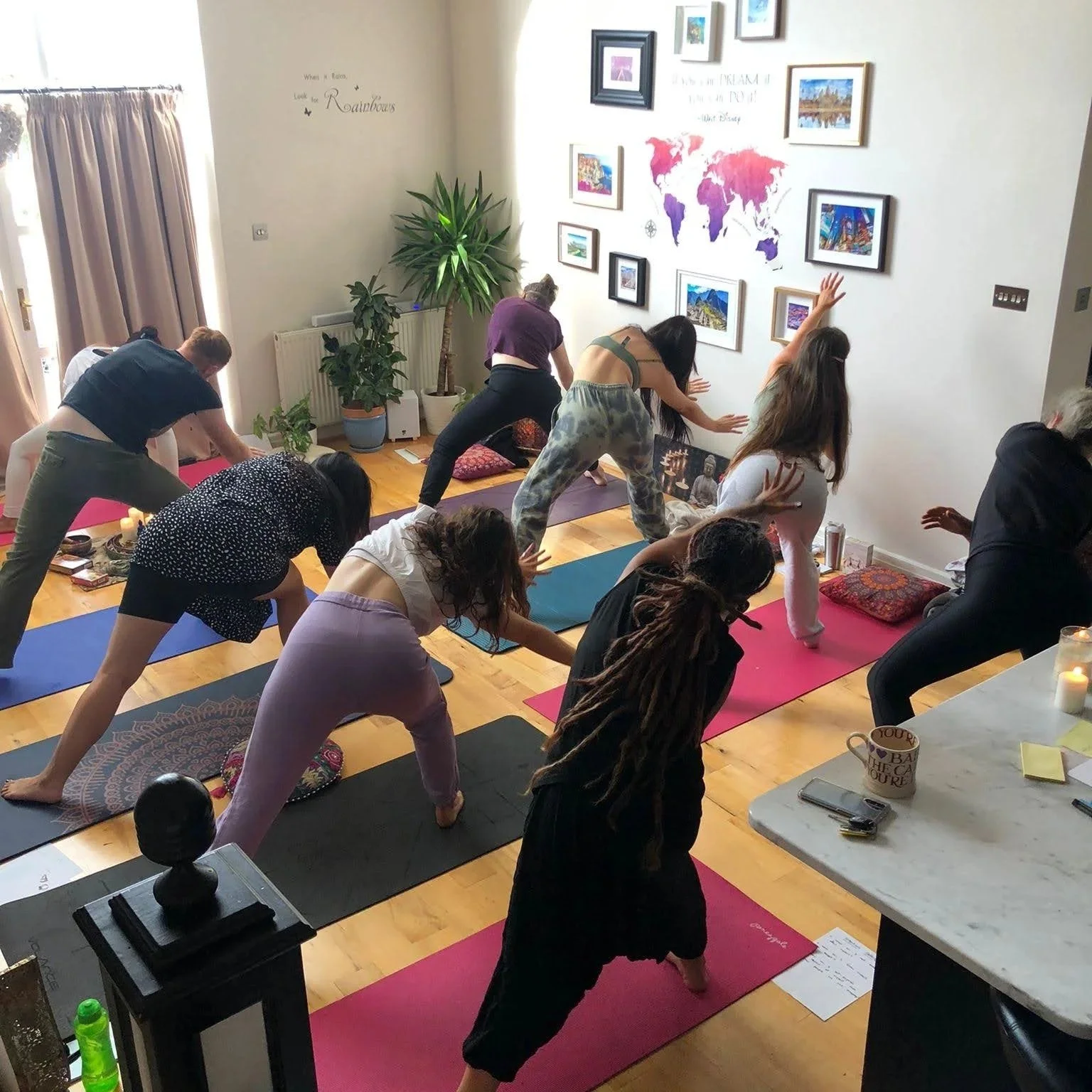 People practicing yoga in a spacious room with wooden floors, light-colored walls, and framed artwork. They are on yoga mats, performing a side stretch pose, and are dressed in comfortable workout clothing. There is a large window with curtains on the left and a wall decorated with a world map and framed pictures.