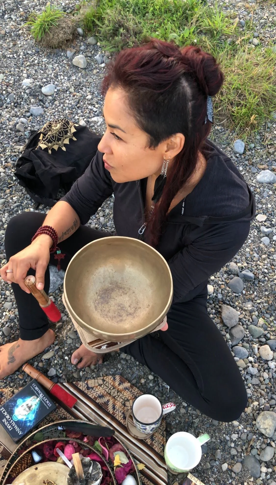 A woman sitting outdoors on a rocky ground holding a singing bowl, with a black backpack, a book, a coffee mug, and a decorative cloth with flowers and candles nearby.