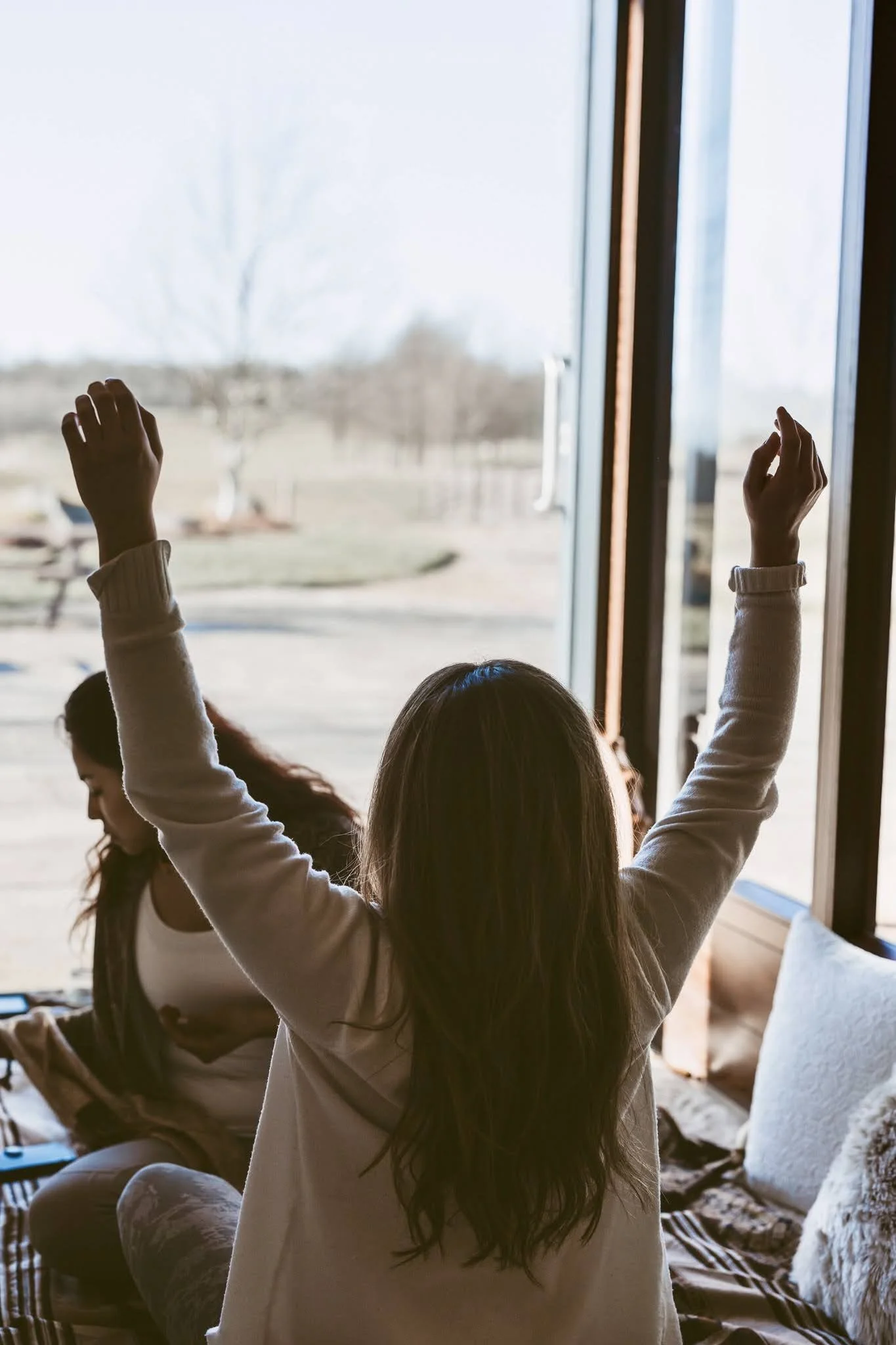 Person with long hair raising hands sitting inside near a large window, with another person looking at a phone nearby.
