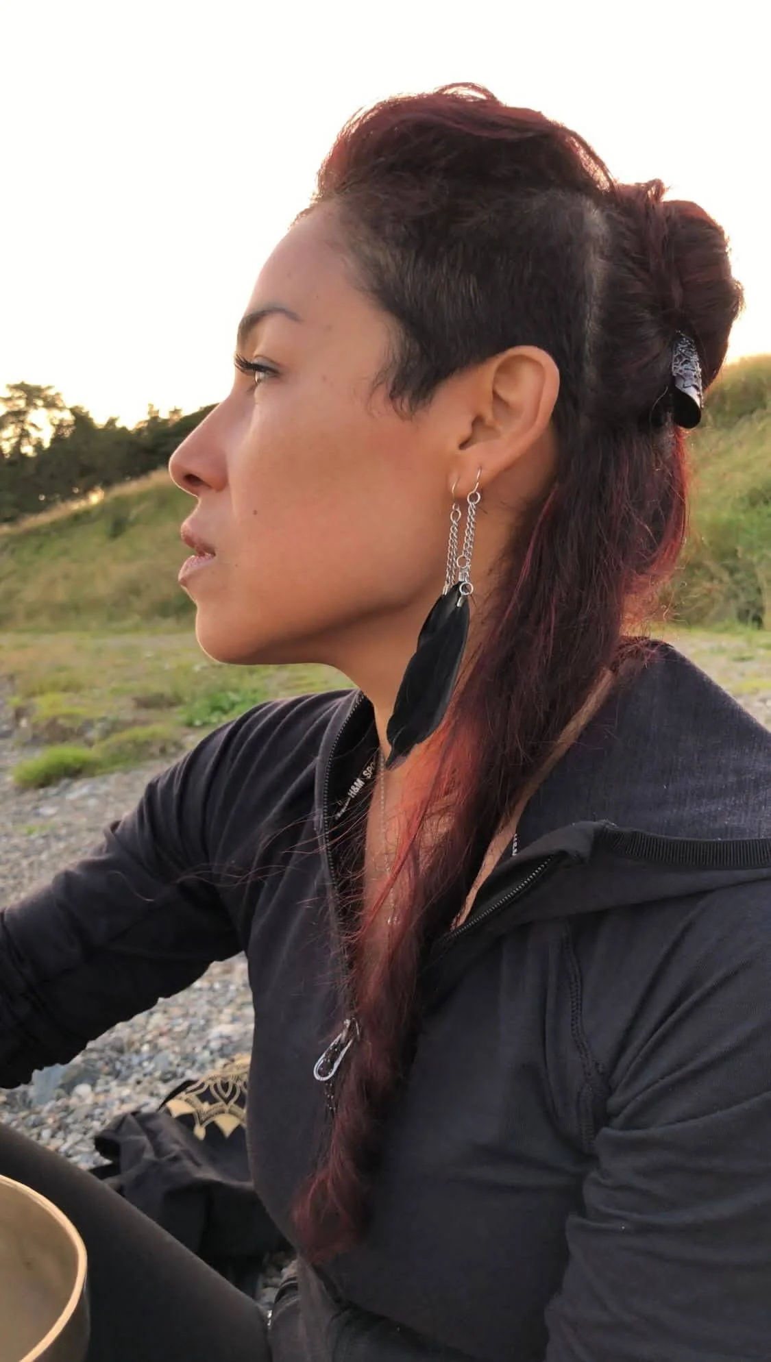 Profile of a woman with elaborate earrings, wearing a black jacket, with a natural outdoor background. She has dark reddish hair styled back, and the photo appears to be taken at sunset.