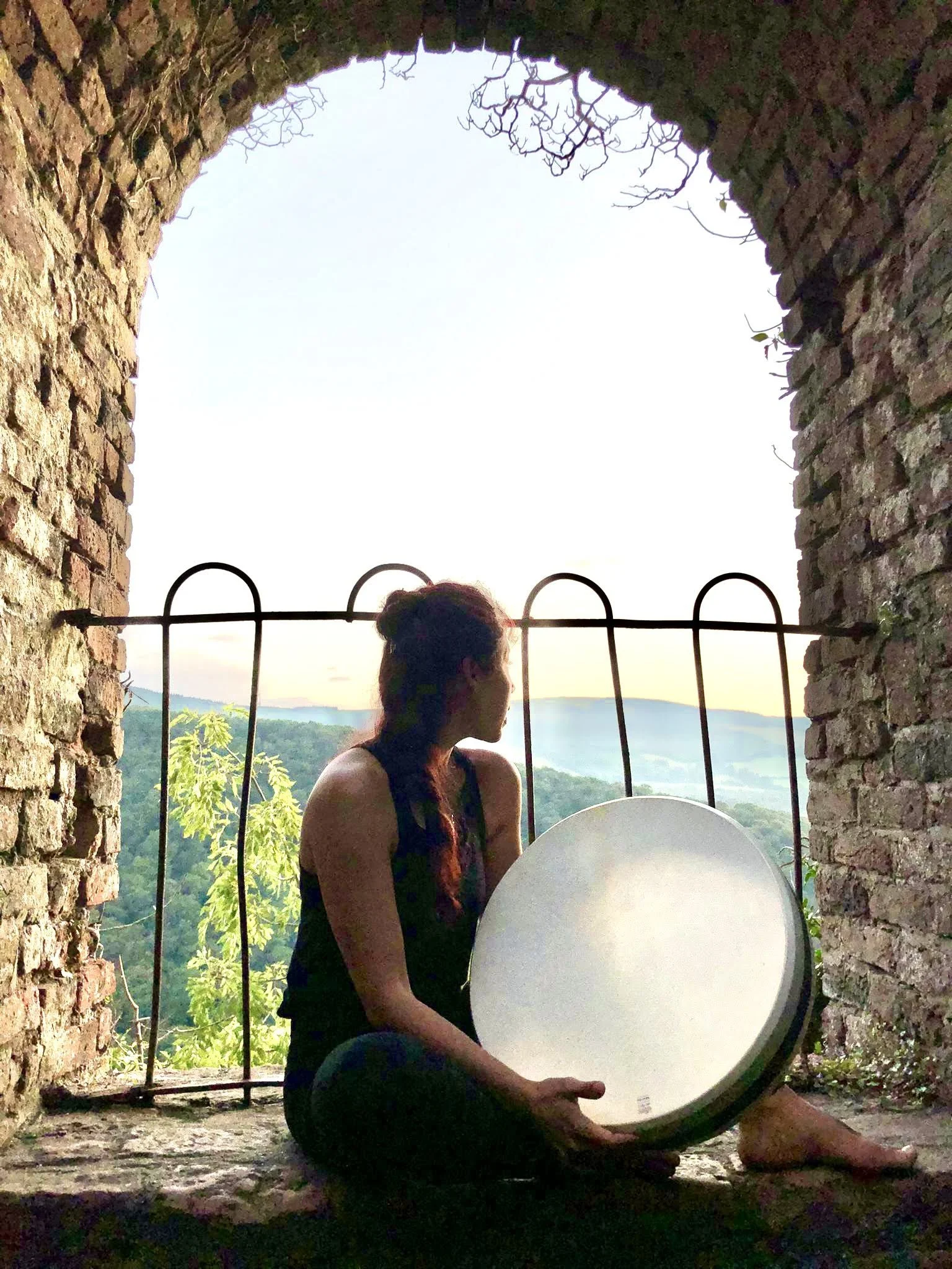 A woman sitting on a stone ledge inside a brick archway, holding a large round drum, looking out at a scenic view of green hills and trees during sunset.