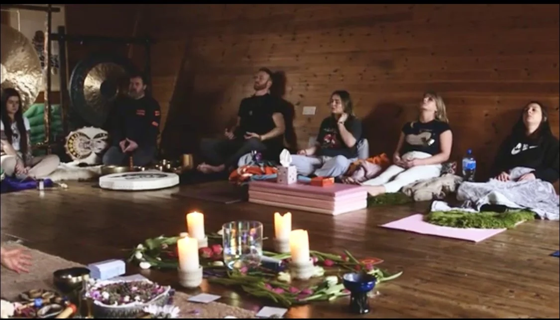Group of people sitting cross-legged on the floor in a wooden room, participating in a meditation or spiritual ceremony, with candles and flowers arranged on the floor in front of them.