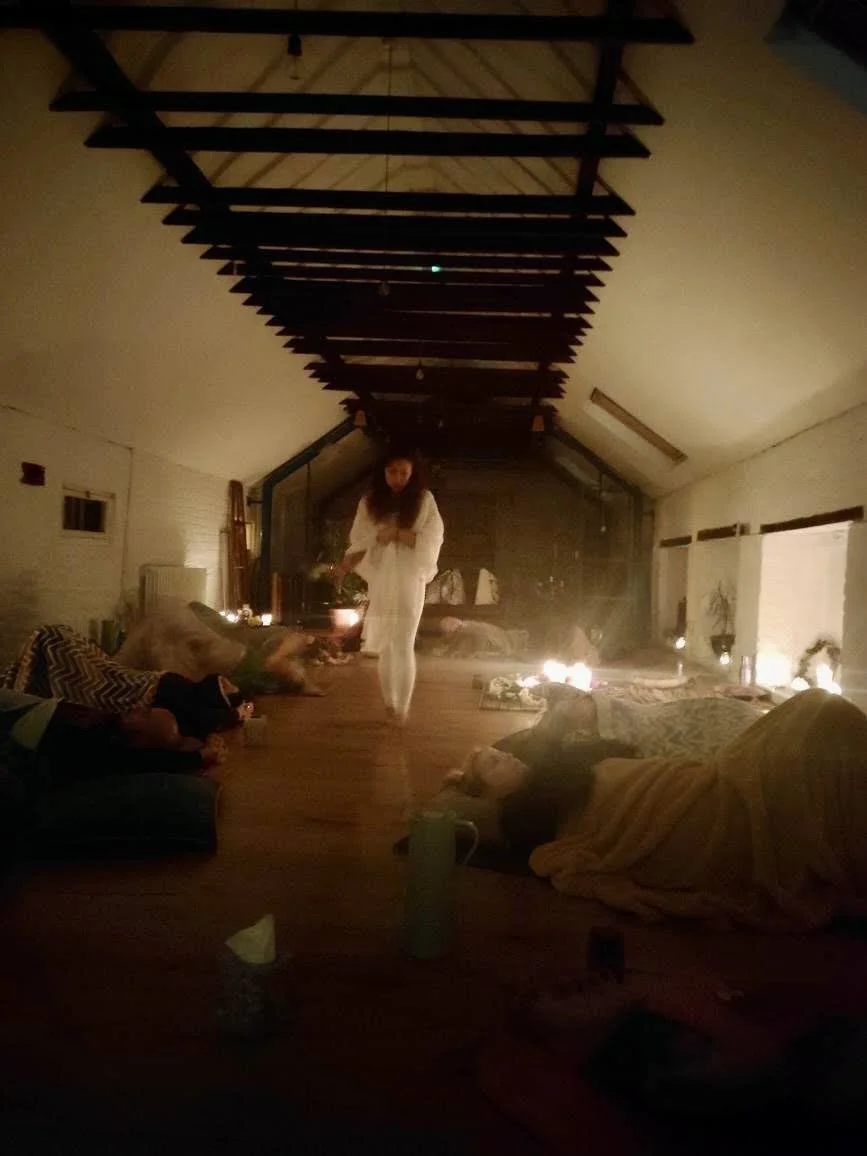 A cozy, dimly lit room with a sloped ceiling and exposed beams, decorated with candles and blankets, where several people are resting or lying on the floor. A woman in white is walking in the center of the room.