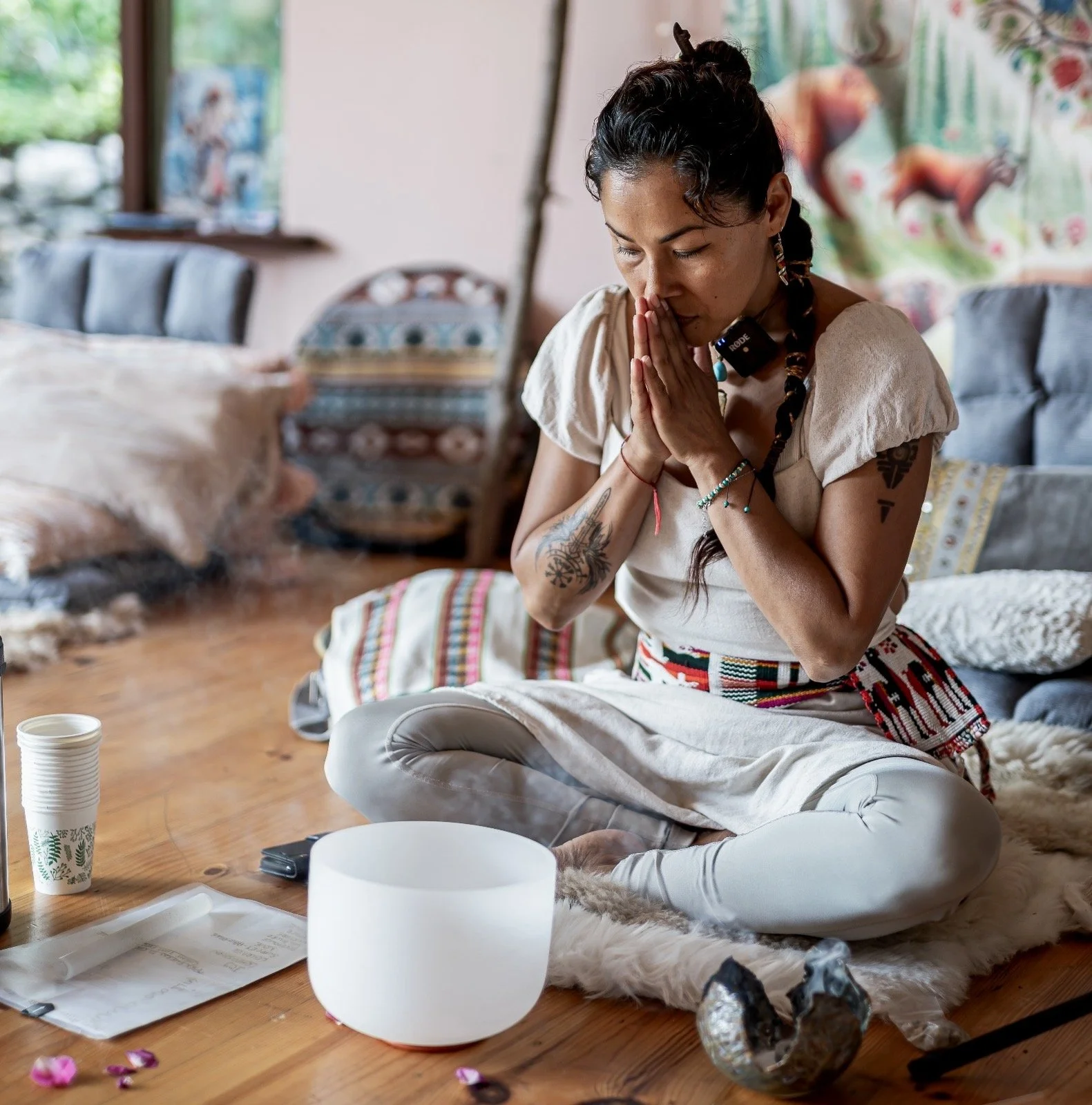 A woman practicing meditation or prayer, sitting cross-legged on a fur rug on a wooden floor, with a singing bowl and various items around her, in a cozy, decorated room with colorful textiles and artwork.
