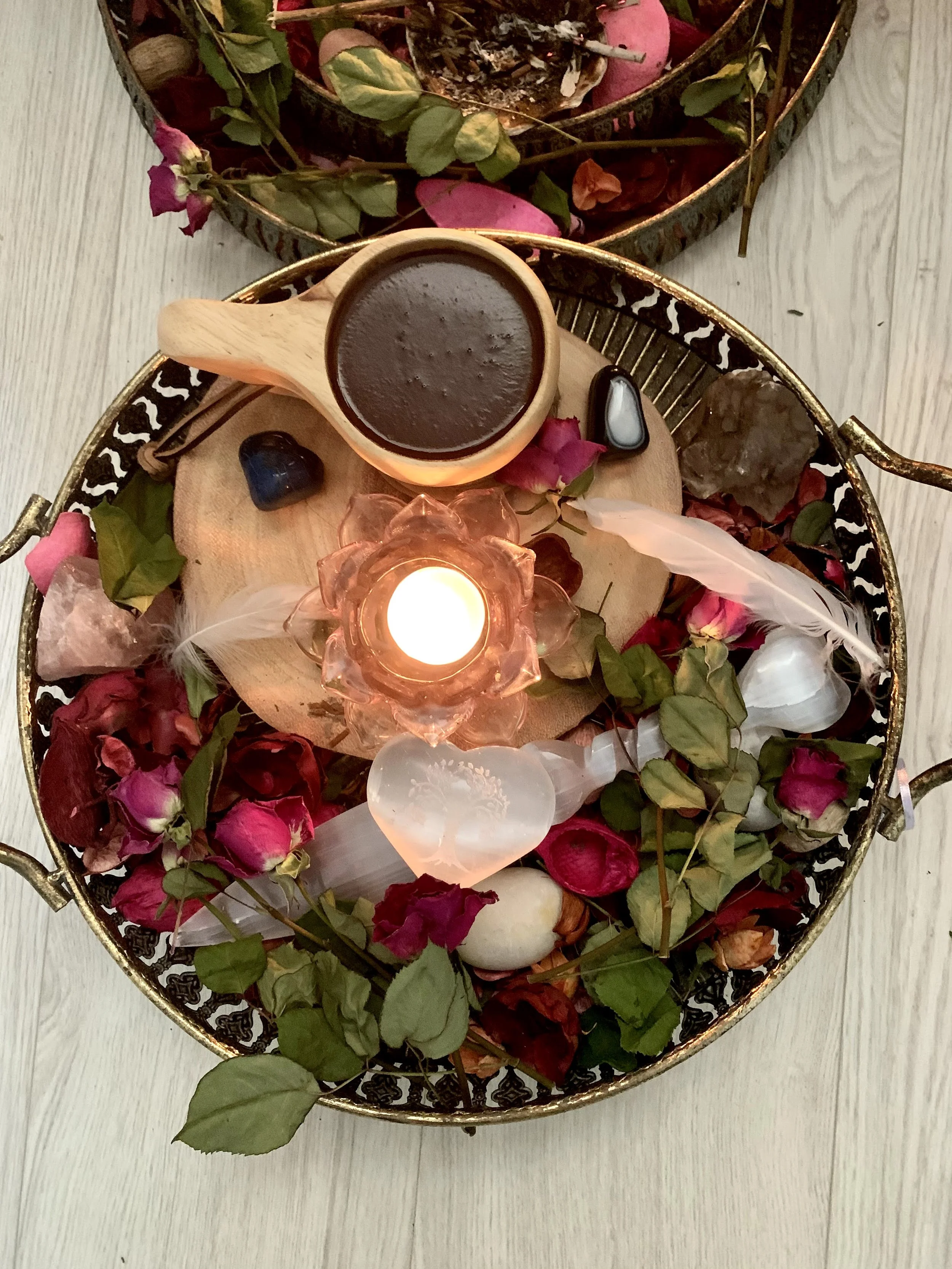 A decorative tray with a candle, a wooden cup of hot chocolate, various crystals, rose petals, and feathers, arranged for a calming or spiritual setting.