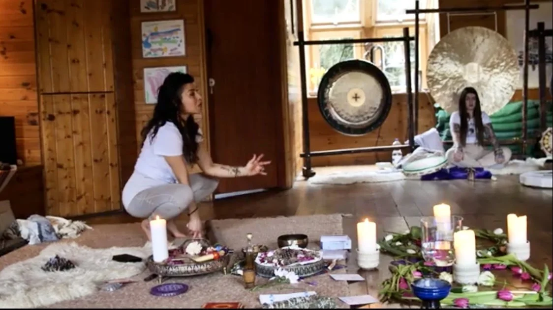 A woman in white clothing is squatting on the floor, making a hand gesture, in a room decorated for a spiritual or meditation session. The floor has lit candles, flowers, and ritual objects, with a large gong and gong stand in the background, and a g