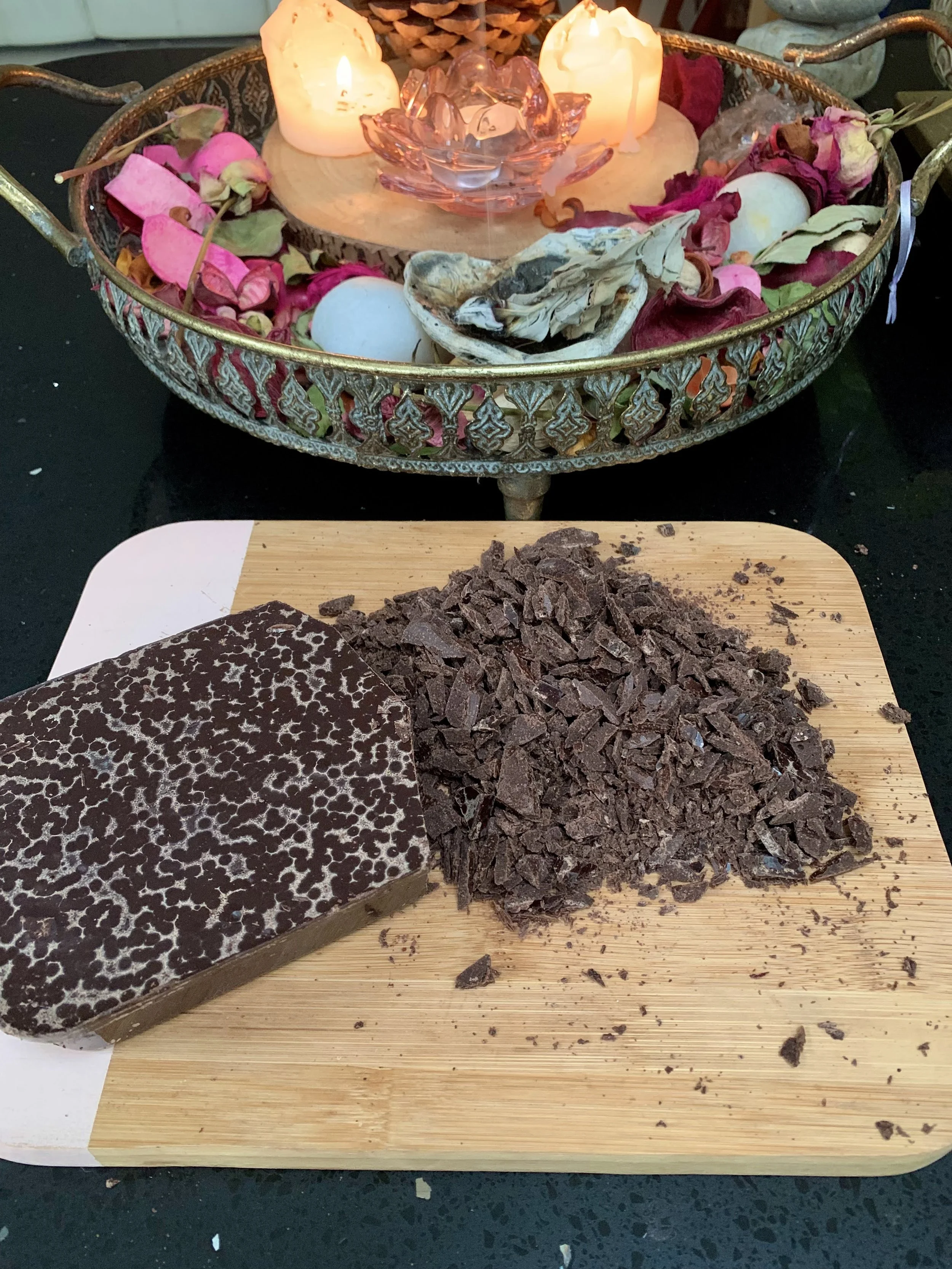 A cutting board with chopped dark chocolate and a chocolate bar. In the background, a decorative tray with candles, dried flowers, and decorative eggs.