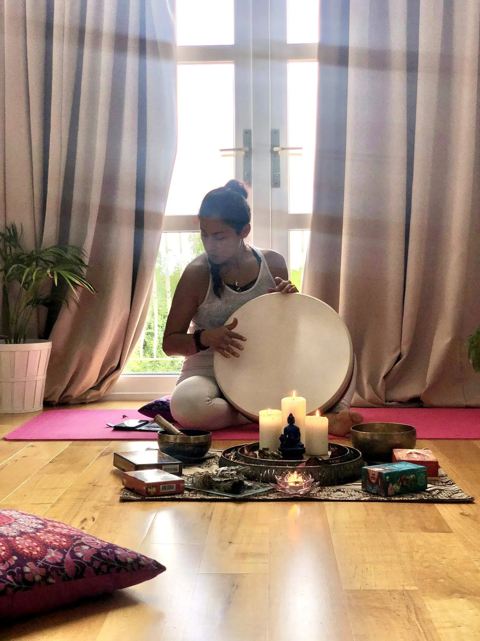 A woman sitting on a pink yoga mat indoors, holding a large circular drum, near candles and spiritual items on a decorative cloth, with a window and curtains behind her.
