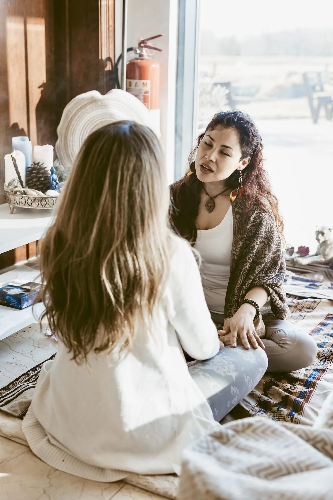 Two women sitting on the floor engaged in conversation, with decorative candles, a pinecone, and jewelry around them, near a large window letting in natural light.