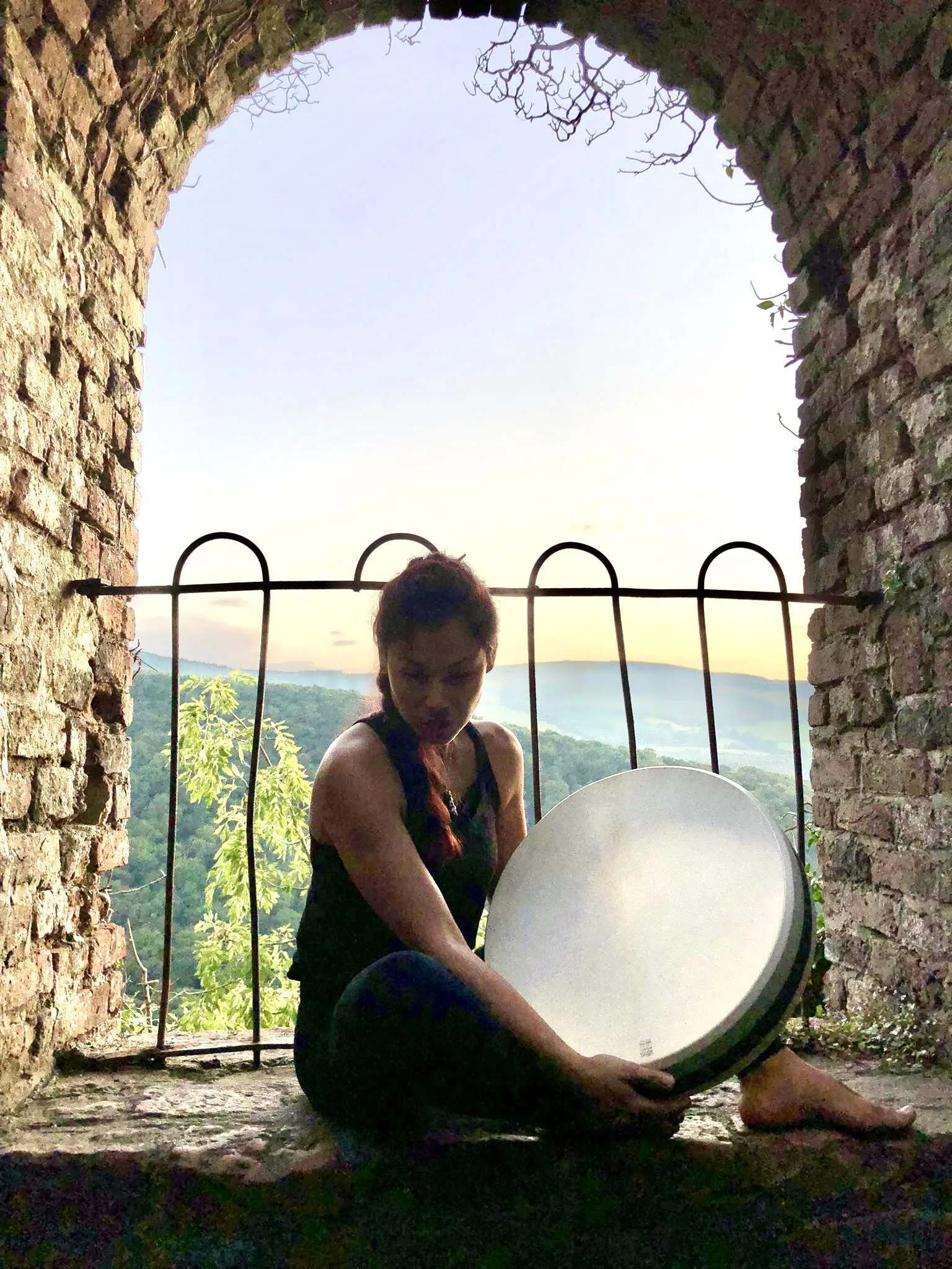 A woman sitting against a brick wall holds a tambourine, with a scenic view of trees and hills through an arched stone window behind her at sunset.