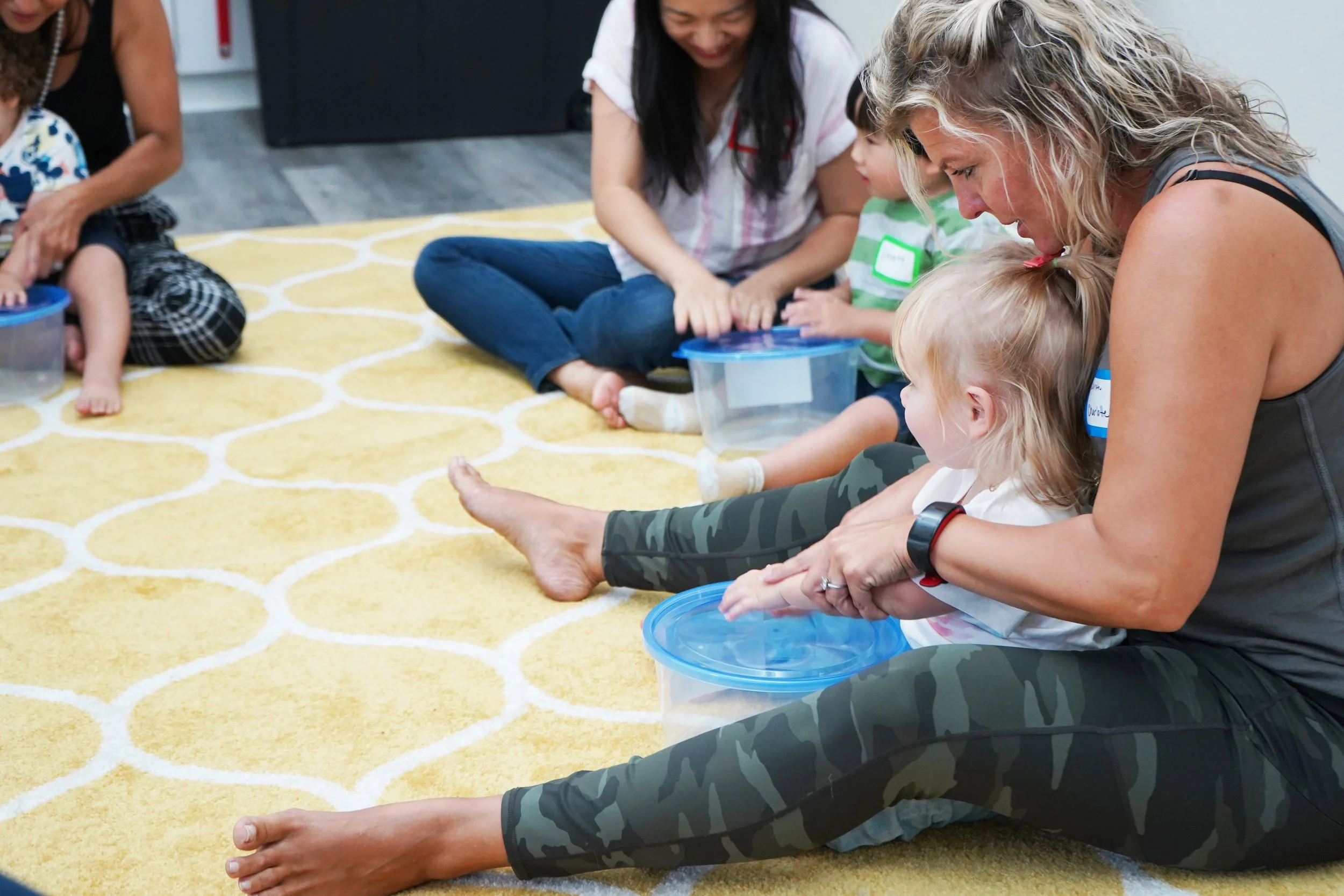 Adults and children sitting on a yellow patterned rug, engaging in a water activity with small blue containers, in an indoor setting.