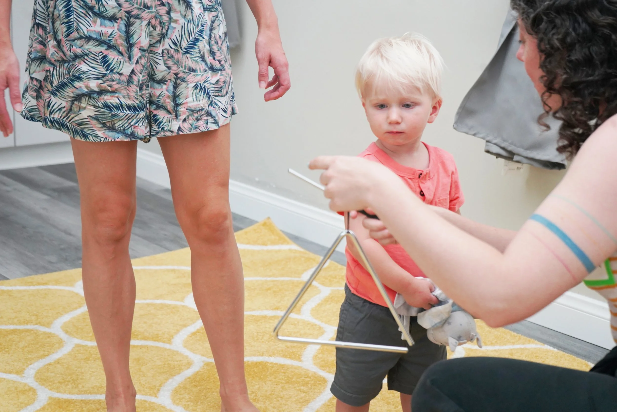 Young boy receiving a triangle-shaped gyroscope from a woman, with another woman standing nearby in a room with a yellow patterned rug.