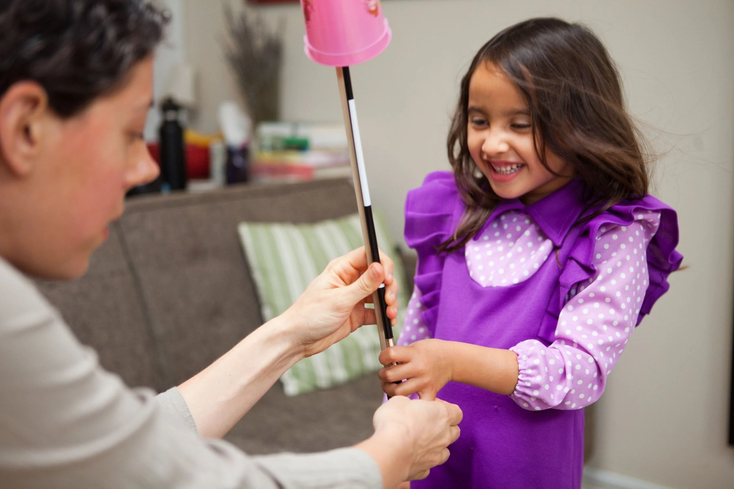 A young girl in a purple dress with ruffles and polka dots on the collar and sleeves smiling as she holds a large pink lollipop, with an adult woman helping her hold the stick.