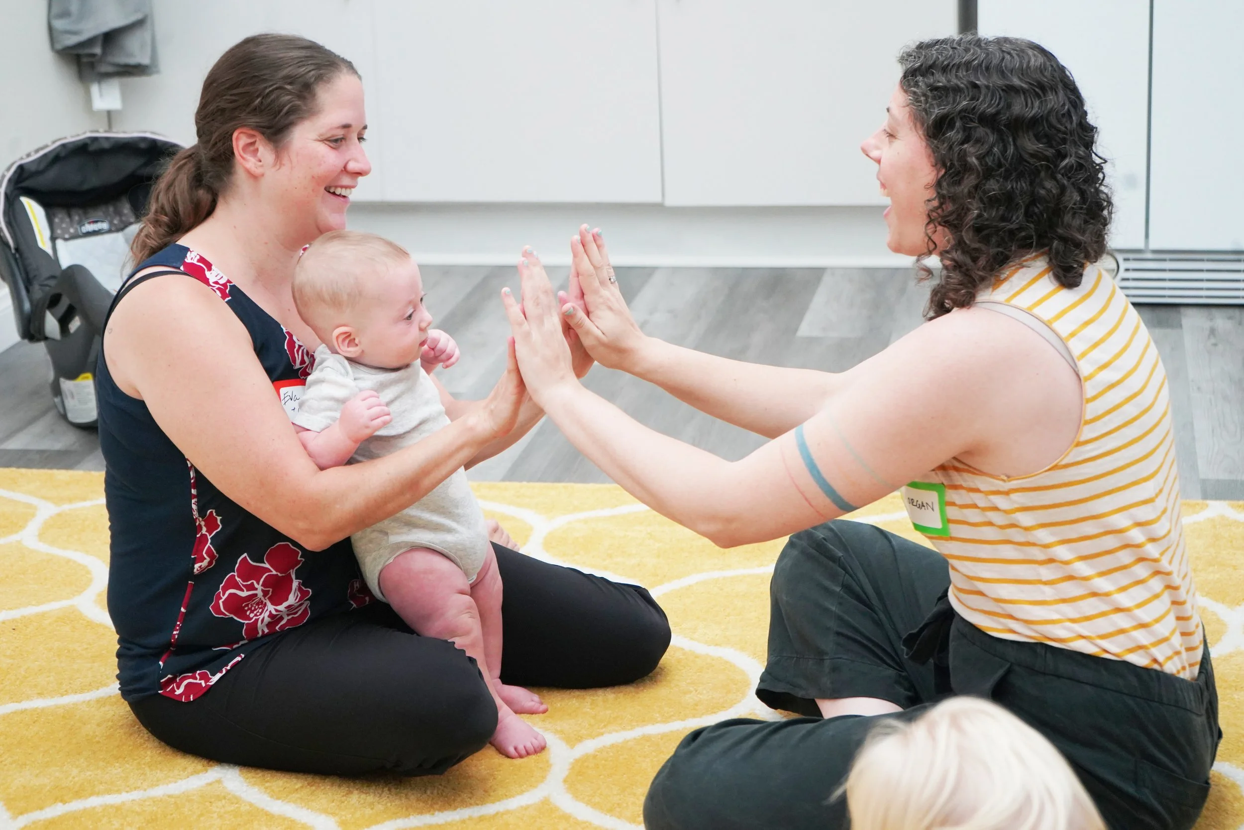 Two women playing finger-touch game with an infant in a room with a yellow carpet and nearby backpack.