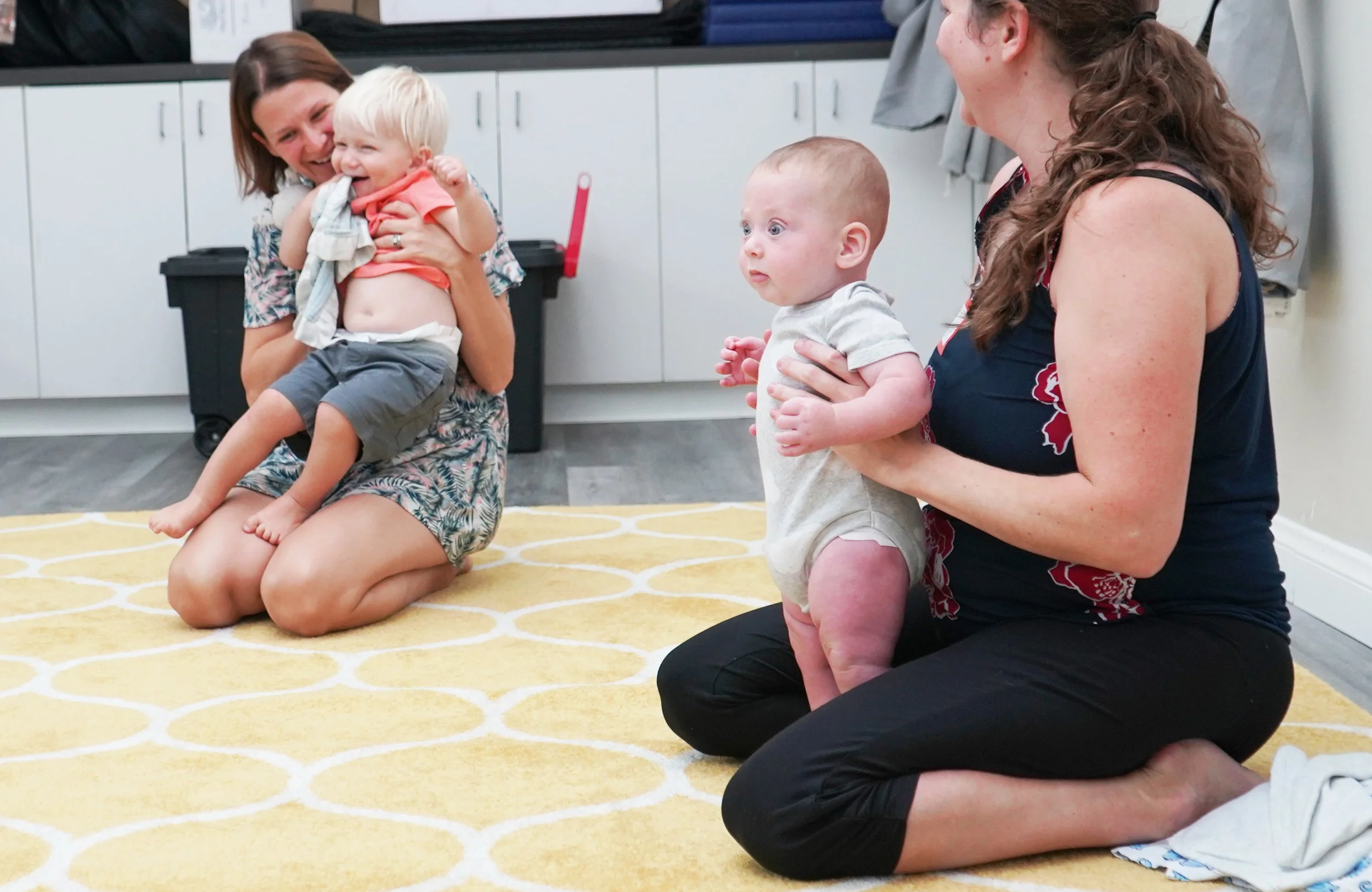 Two women and two small children in a room with white cabinets and a yellow patterned rug. One woman is kneeling on the floor, holding a laughing child with blonde hair and a red shirt. The other woman is sitting on her knees, holding a baby with light skin and chubby cheeks, wearing a light-colored onesie. They are engaging in a playful, joyful activity together.