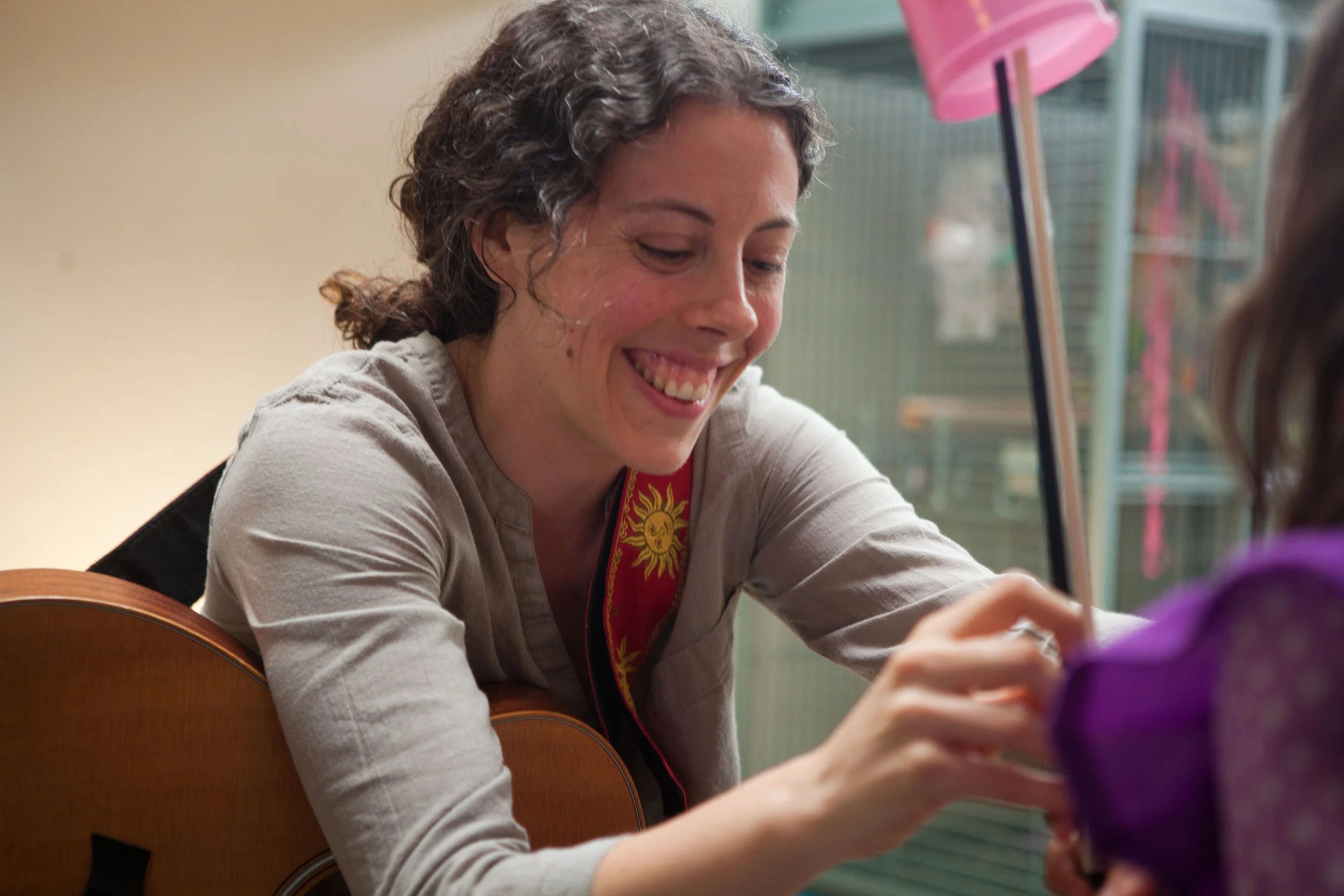 A woman with curly hair smiling while playing a guitar and drawing with a colorful marker on a child's arm or hand.