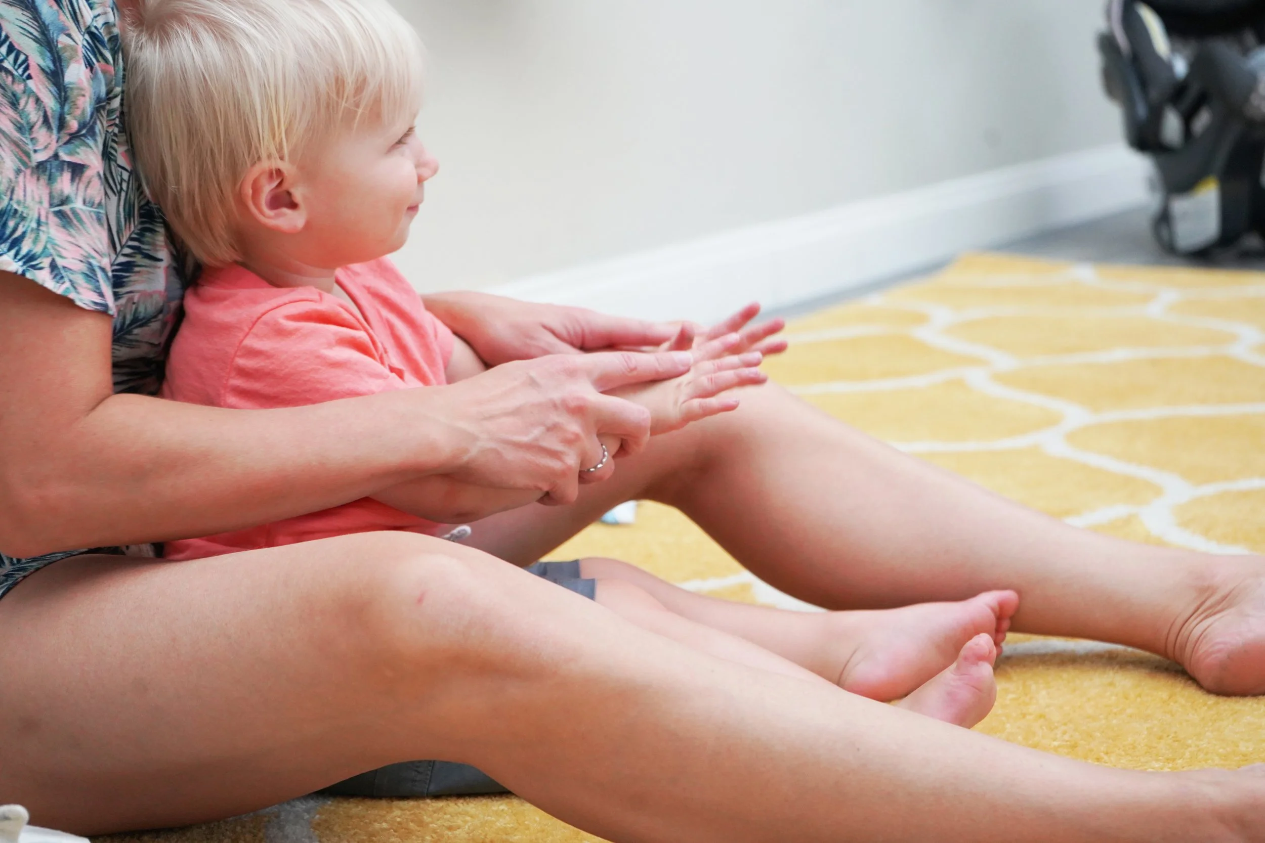A woman helping a young girl stretch her legs on a yellow patterned carpet in a room, with the woman's arms assisting the girl and the girl smiling and looking forward.