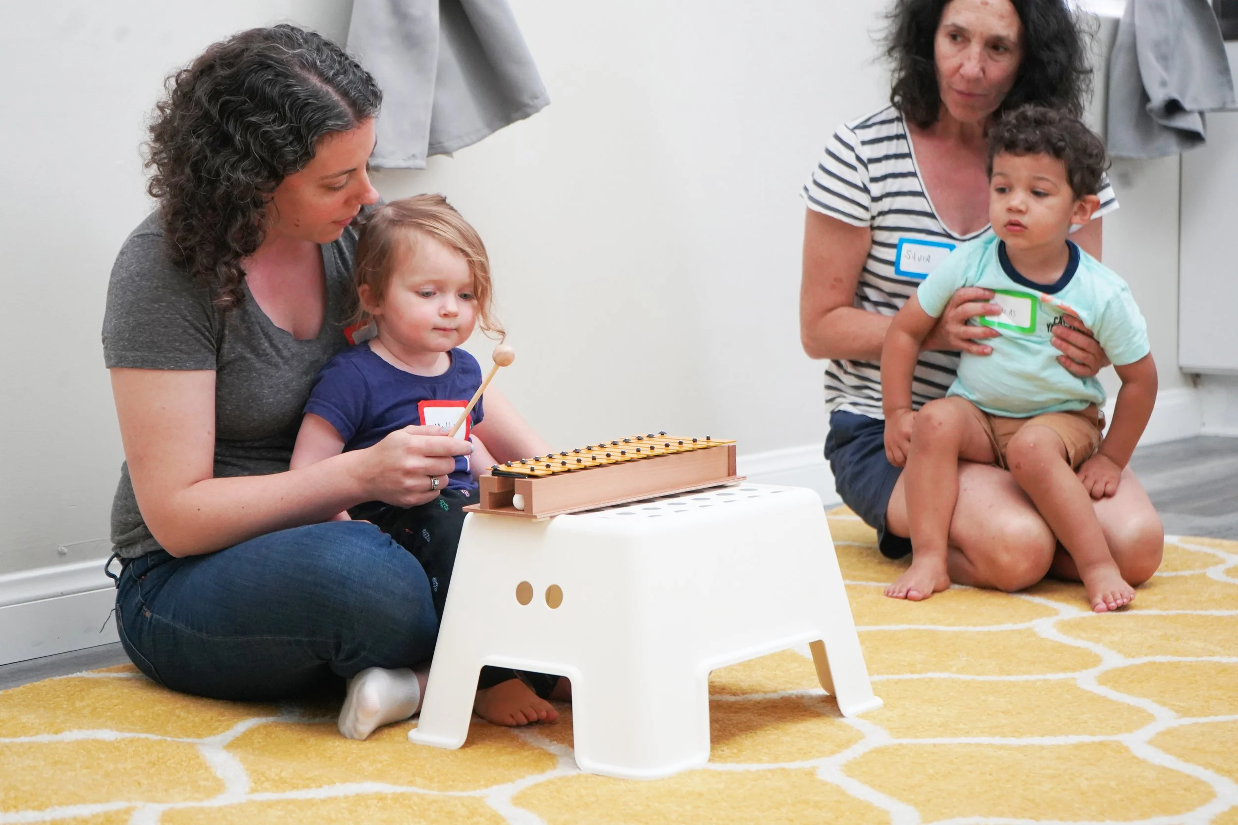 Two women and two children sitting on a yellow rug with white patterns, watching a girl playing a wooden xylophone.