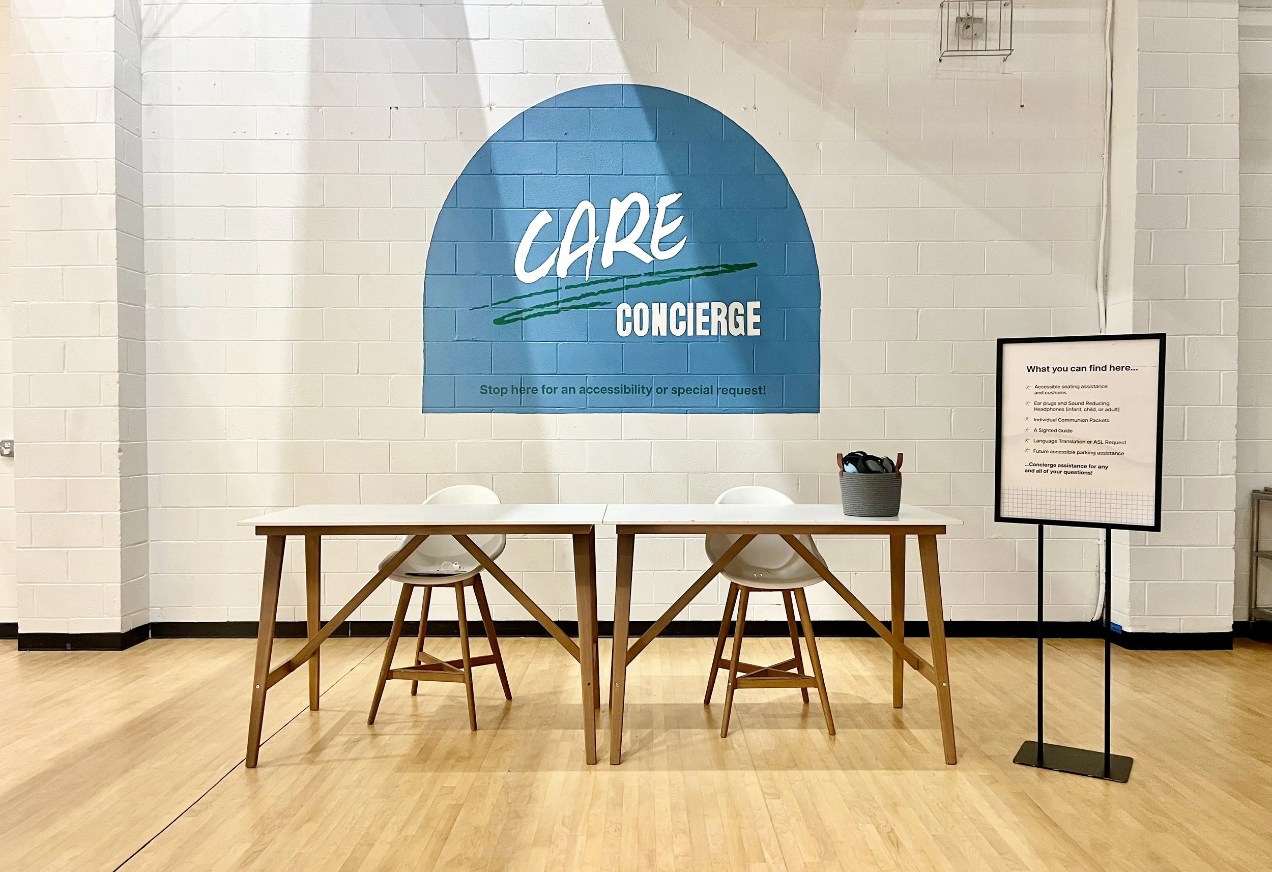 A reception area with a large blue circle wall decoration that reads 'CARE CONCIERGE'. There is a table with two chairs, a basket with items, and a framed sign listing services offered, such as accessibility assistance, hearing aid support, and language translation.