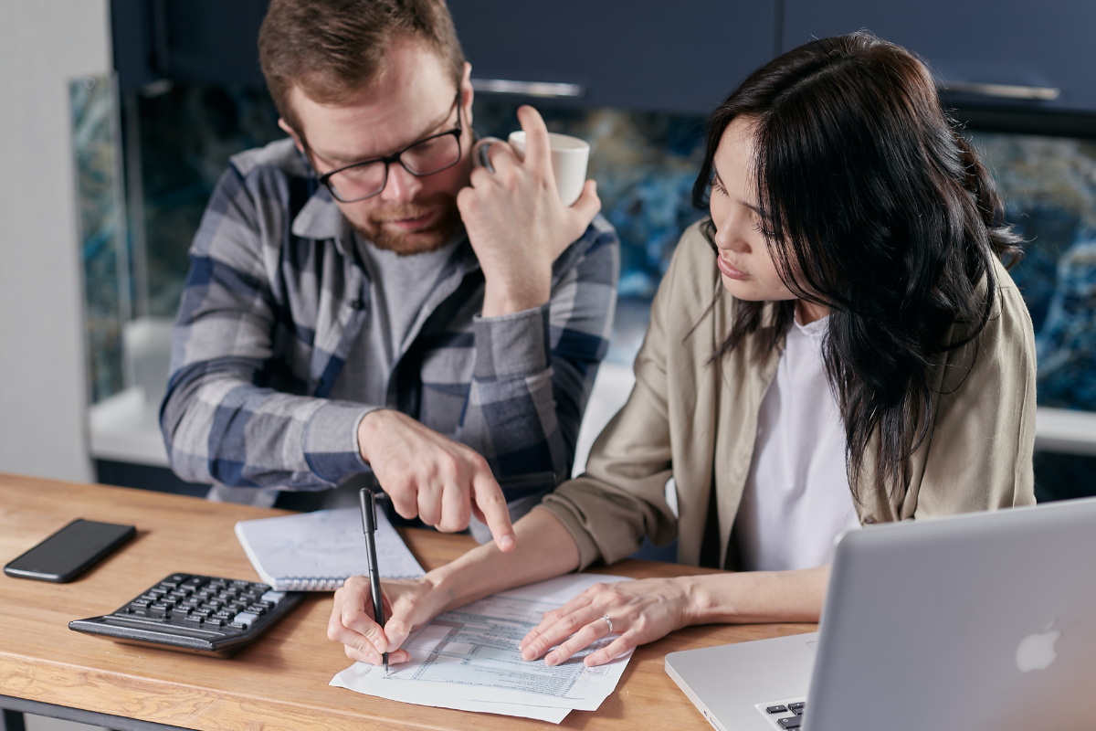 Man and woman working together at a desk, reviewing documents, with a calculator, smartphone, and laptop on the desk.