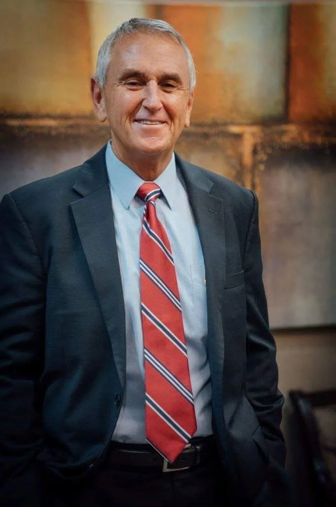 A smiling older man in a business suit with a striped red, white, and blue tie, standing indoors with an abstract background.