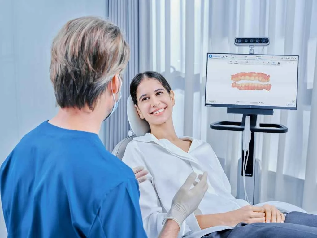 A woman patient smiling while talking to a dentist in a dental clinic with a computer screen displaying a digital image of teeth.
