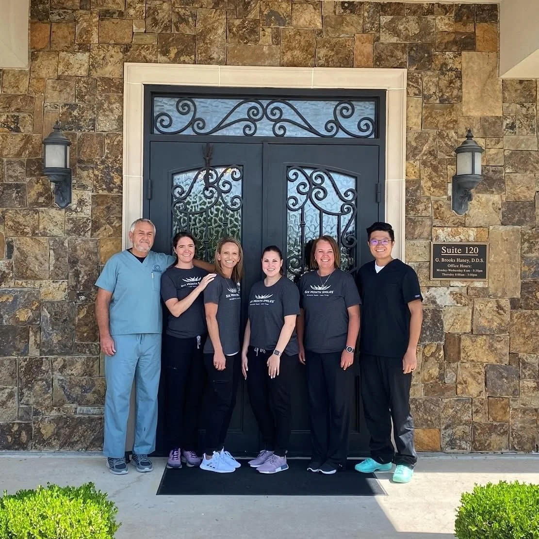 Group of five people standing in front of a stone wall and decorative door, posing for a photo. They are smiling and wearing casual scrubs and t-shirts.