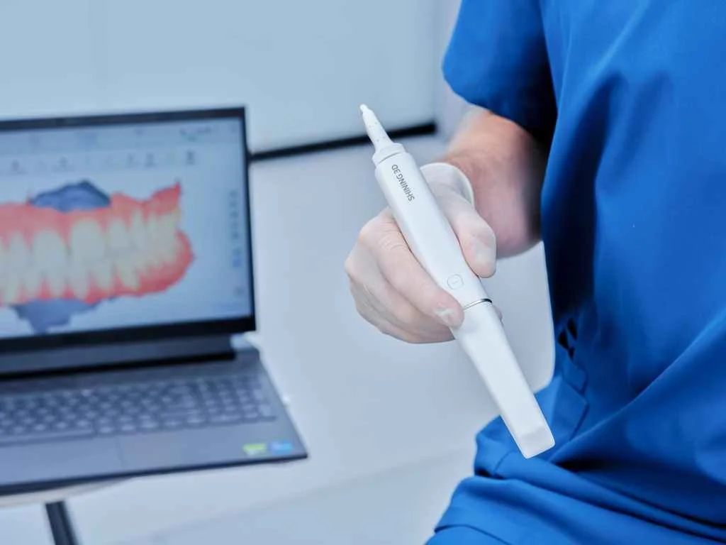 A person in blue scrubs holding a dental scanning pen next to a laptop displaying a 3D dental scan.