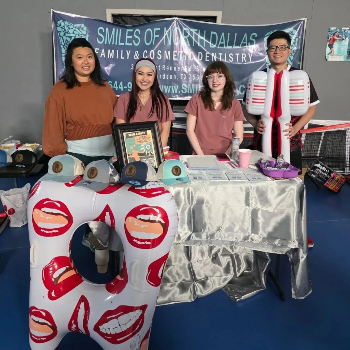 A group of four people standing behind a table at a promotional event for smiles of North Dallas family and cosmetic dentistry. The table is covered with a shiny silver tablecloth and displays hats, flyers, a framed poster, and a purple container with smaller items. There is a large inflatable decoration in front of the table with red and pink lips. A banner in the background displays the business name and contact information. One person wears a costume resembling a large dental bottle.