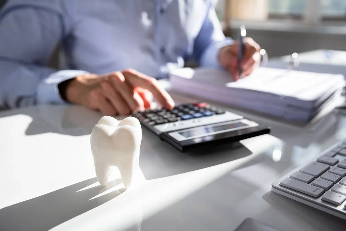 A person in business attire working at a desk with a calculator, a stack of documents, a keyboard, and a model of a tooth on the desk.