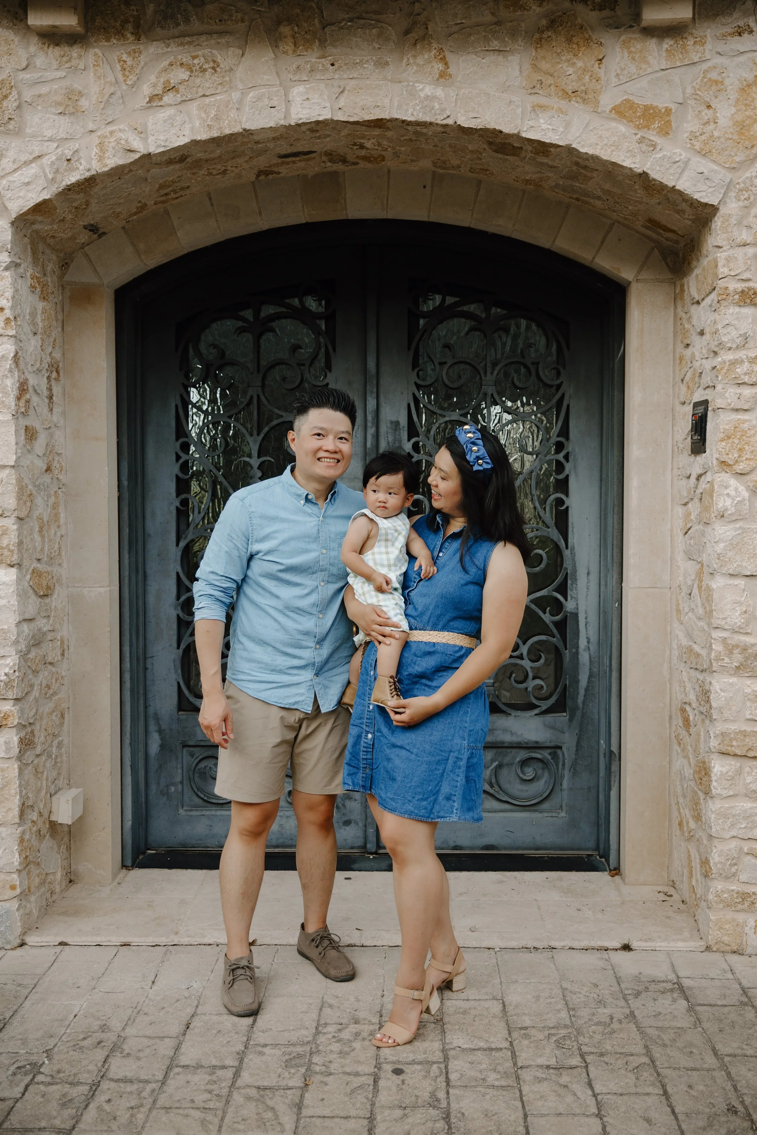 Family of three standing in front of a stone wall and a black metal gate. The man is wearing a light blue shirt and beige shorts, smiling. The woman is wearing a blue dress with a belt and a matching headband, holding a young girl in a light-colored dress. The child is looking at the camera with a neutral expression.