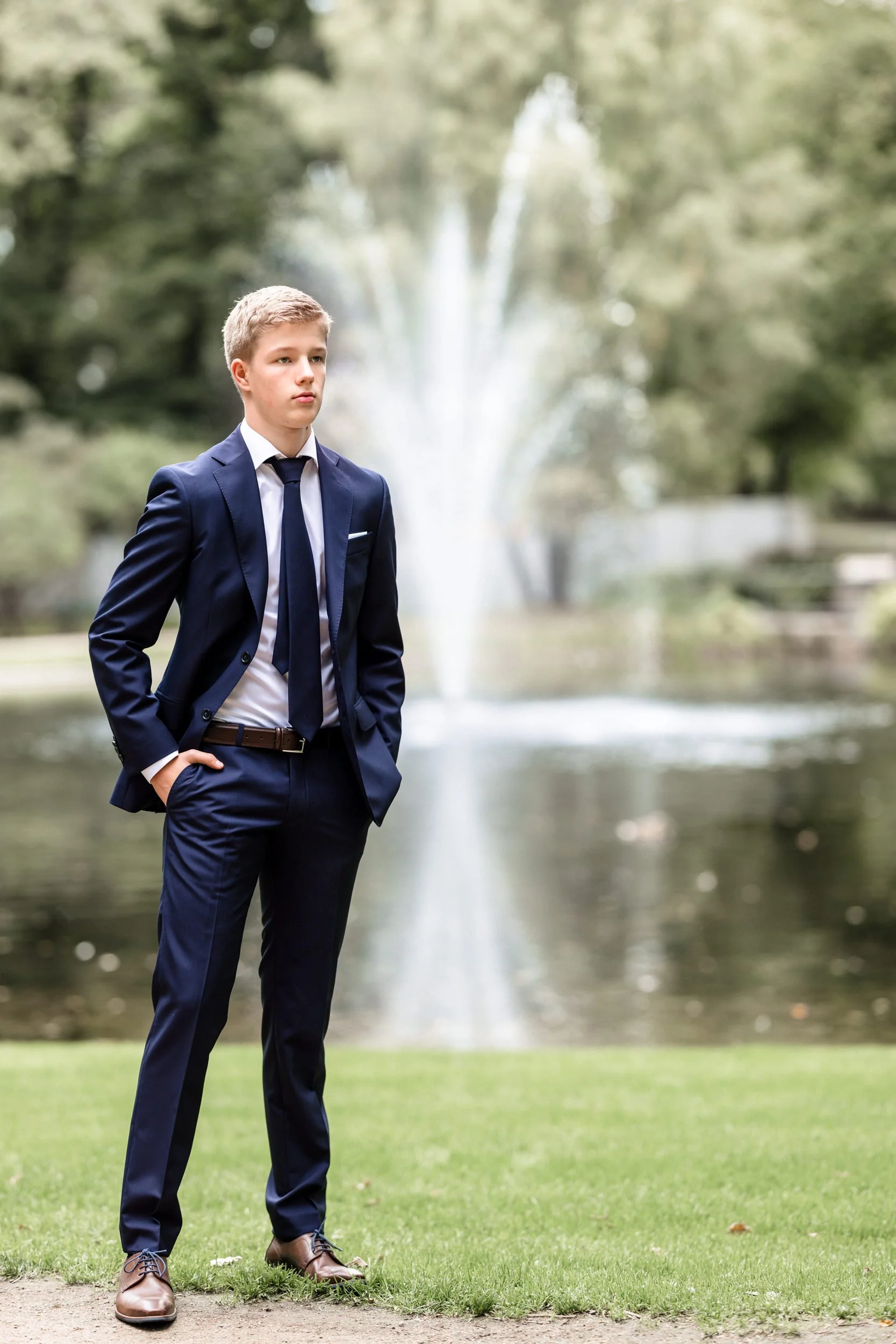 Konfirmant  i en mørkeblå dress står med hendene i lommene foran en fontene i en park.