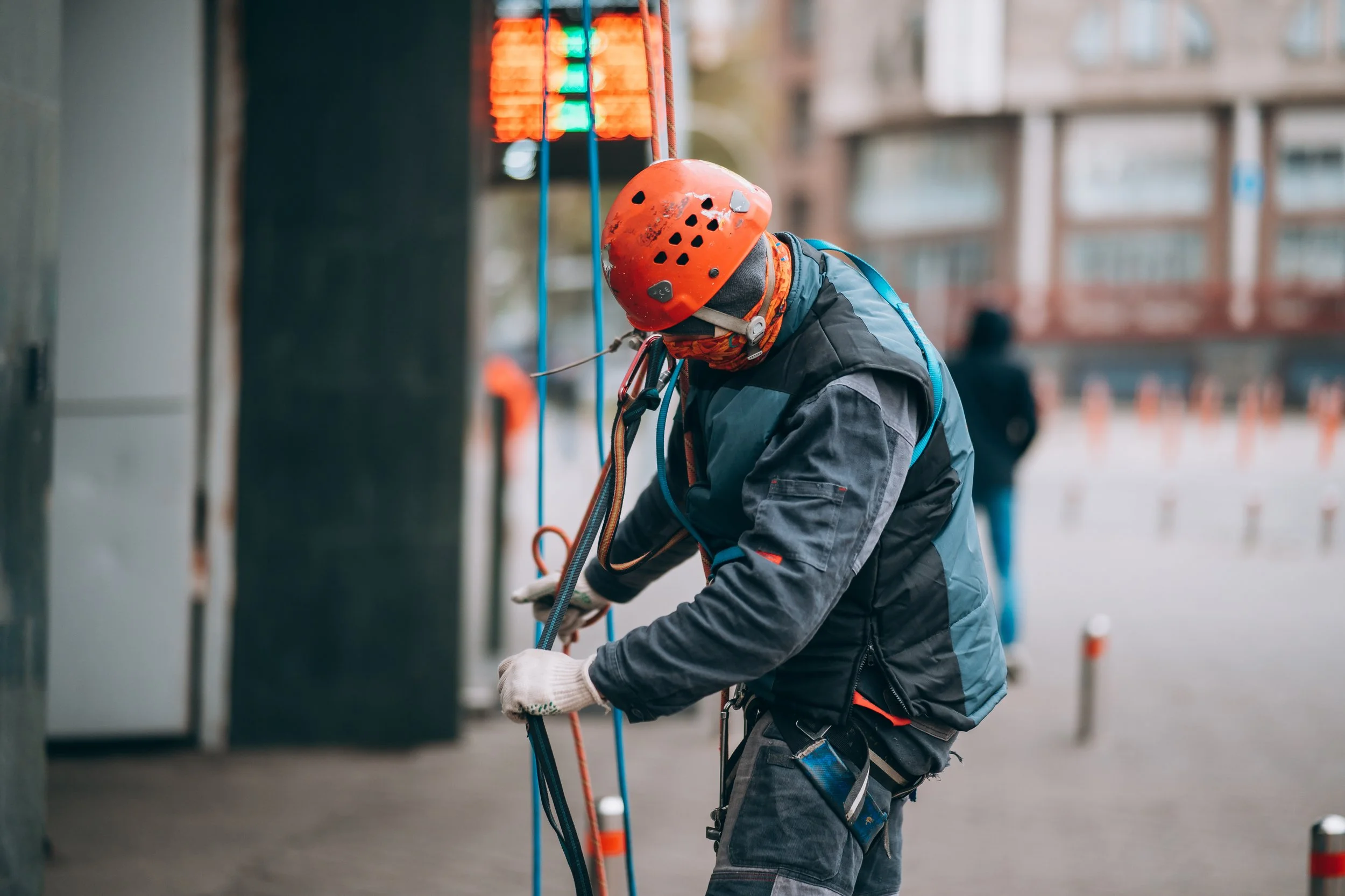 Construction worker wearing safety gear, helmet, and harness, working with blue and orange ropes on a city sidewalk.