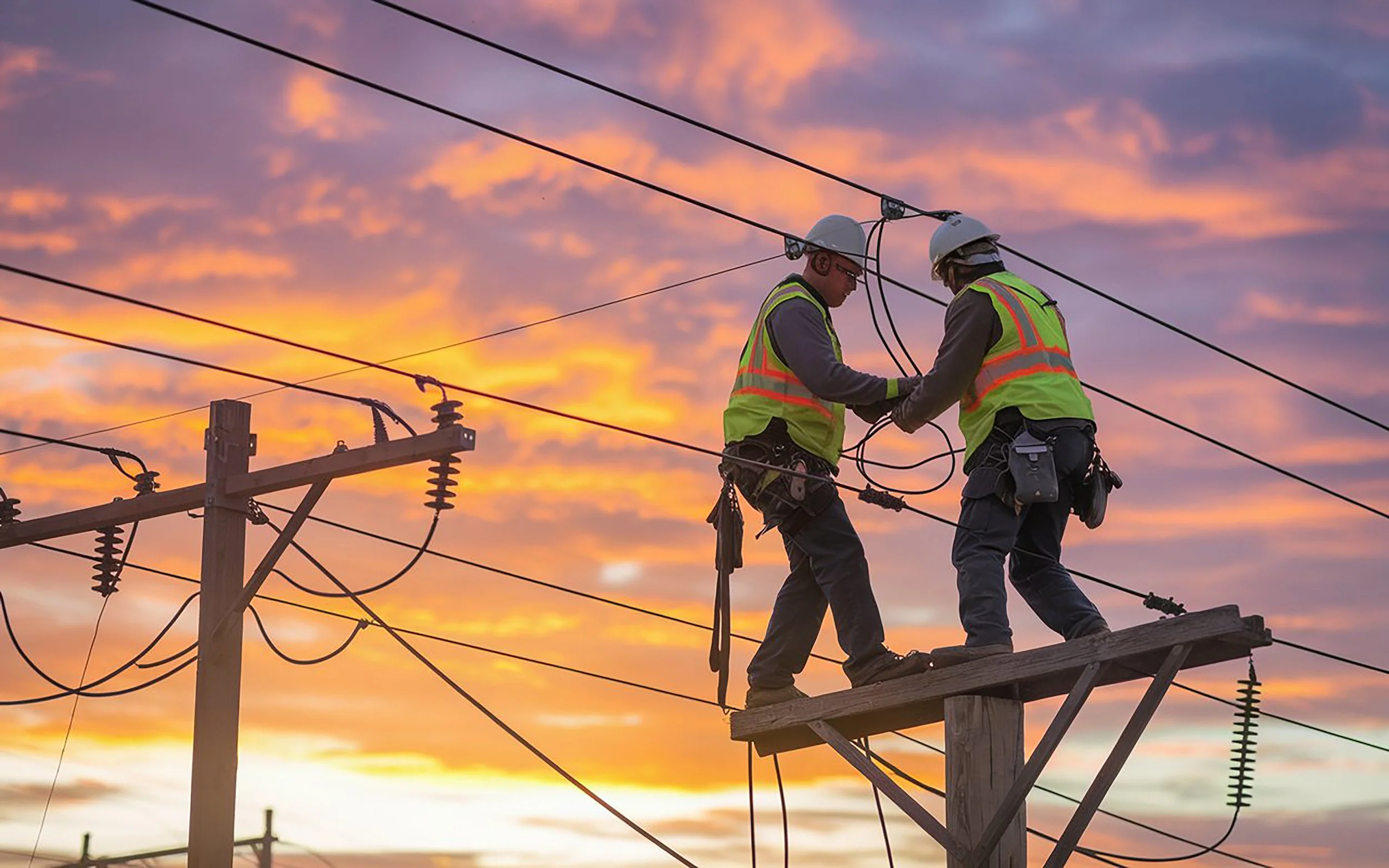 Two electrical workers wearing safety helmets and reflective vests working on power lines during a sunset.