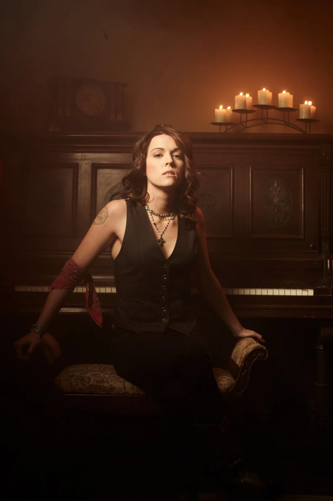 A woman with dark hair and tattoos sitting on a vintage chair in front of a piano, lit by candles on a candelabrum, creating a moody, dimly lit atmosphere.