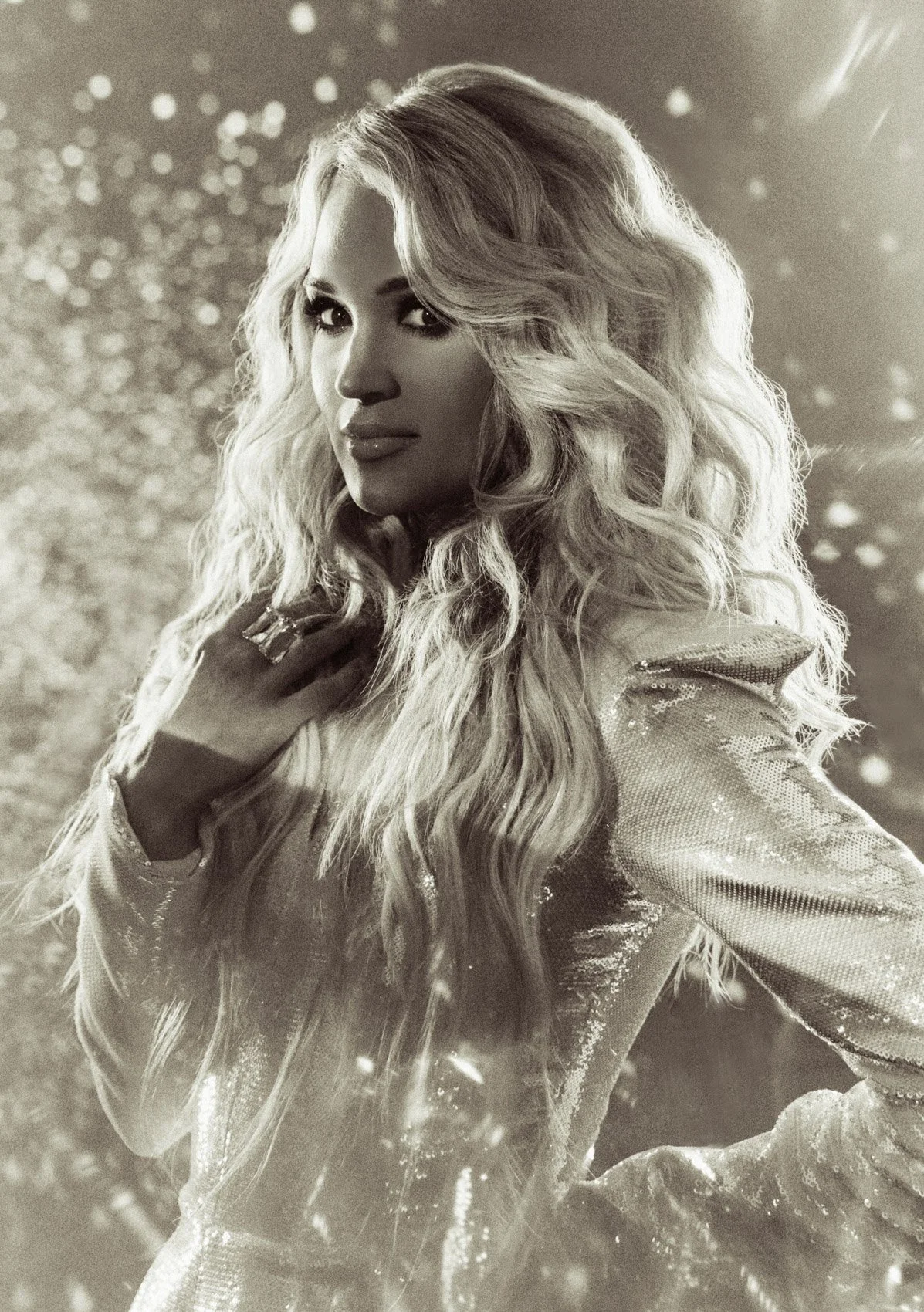 A black and white photo of a woman with long, wavy hair, wearing a shiny, textured jacket, with a sparkly background and light effects.