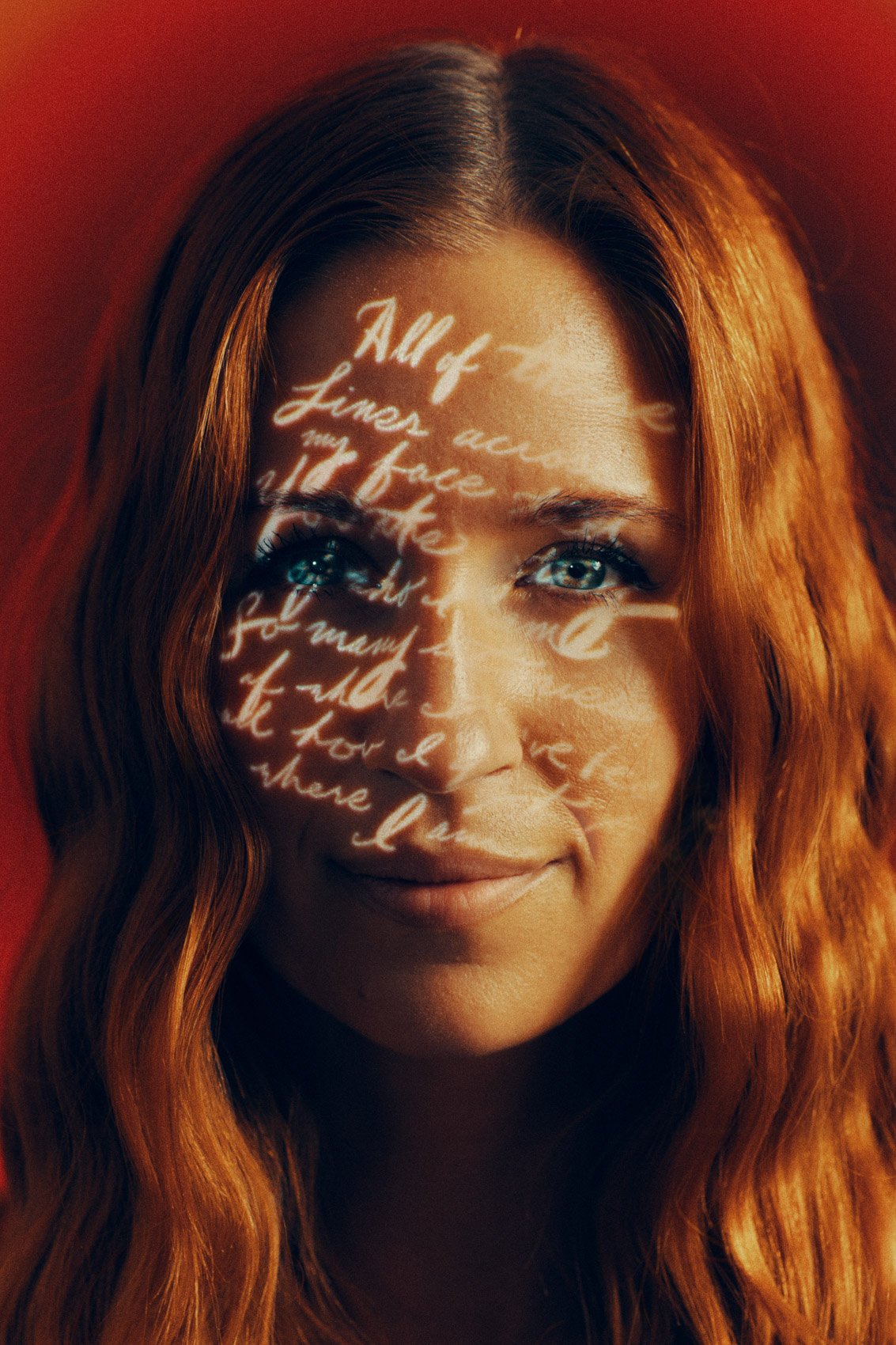 A woman with wavy red hair and blue eyes has a reflection of white handwritten text projected onto her face, with a red background.