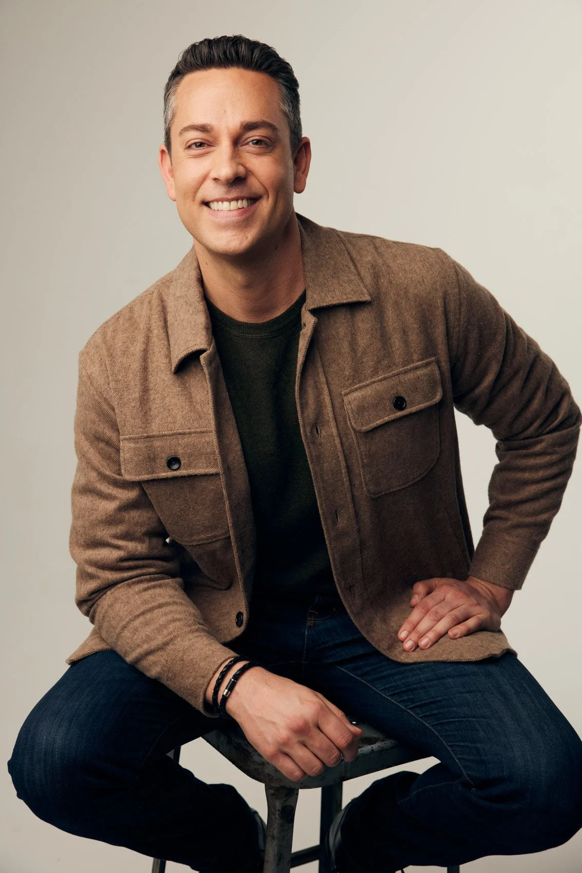 A smiling man is sitting on a stool against a plain light-colored background. He has short dark hair and is wearing a brown jacket over a dark shirt and dark jeans.