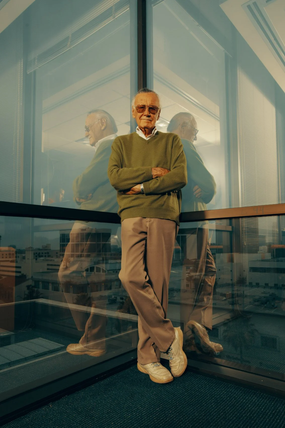 An elderly man with glasses standing with arms crossed next to a glass wall. His reflection is visible in the glass, creating a mirrored effect. The background shows a cityscape through the window.
