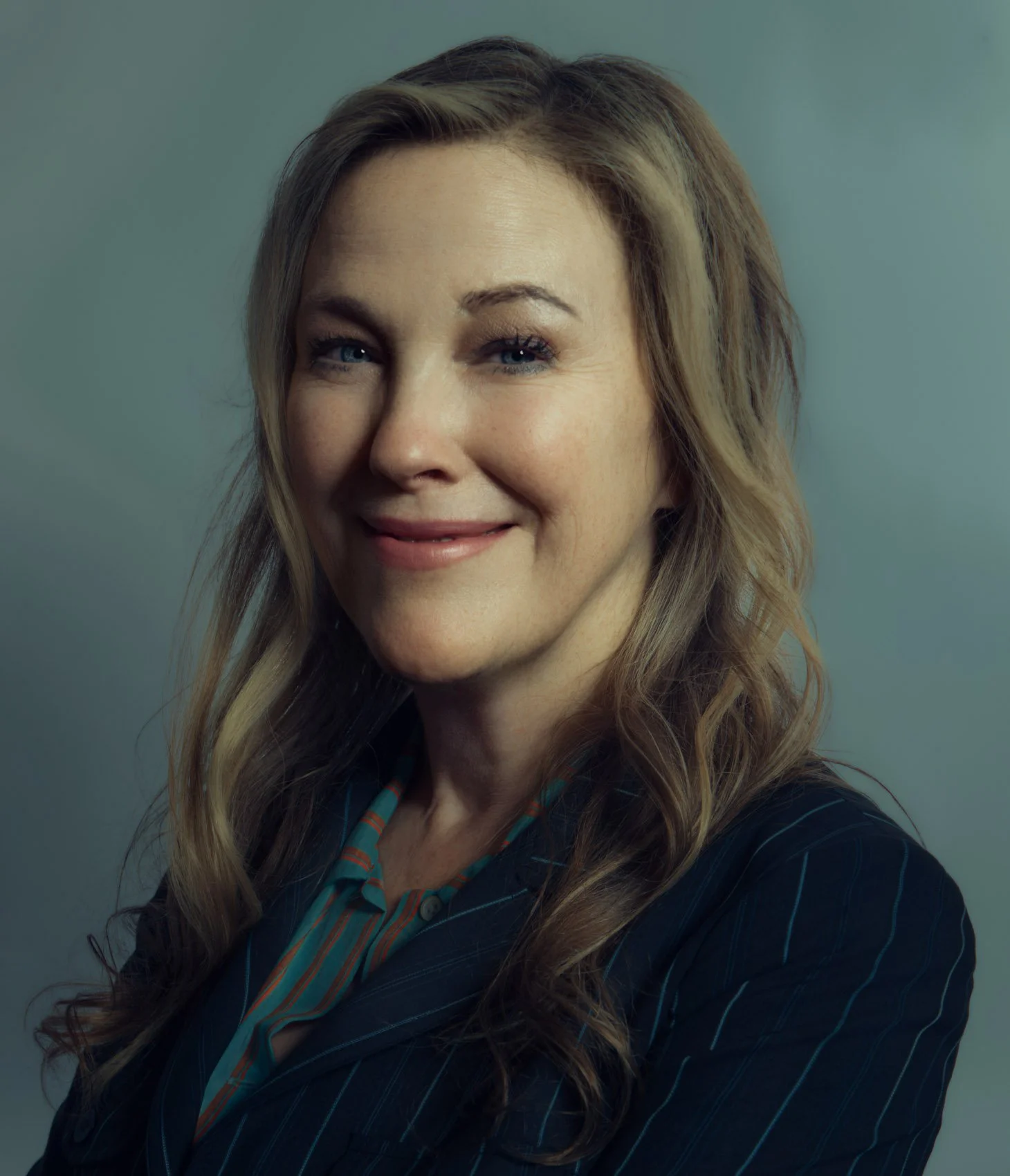 A woman with long, wavy blonde hair wearing a dark pinstripe blazer and a colorful blouse smiling at the camera against a plain background.