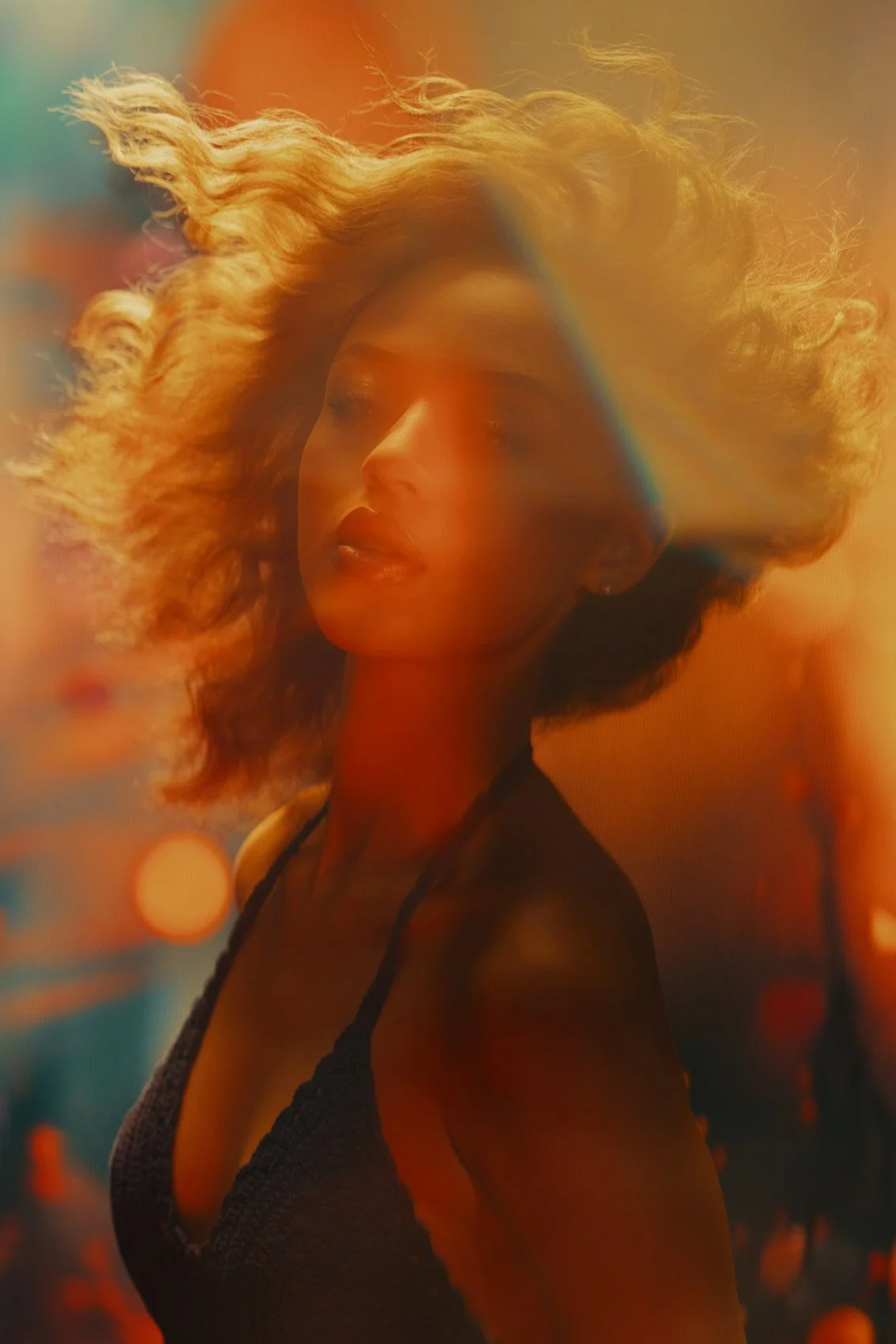A woman with curly blonde hair and dark clothing looking to her right, with soft warm lighting creating a blurred, colorful background and rainbow lens flare.
