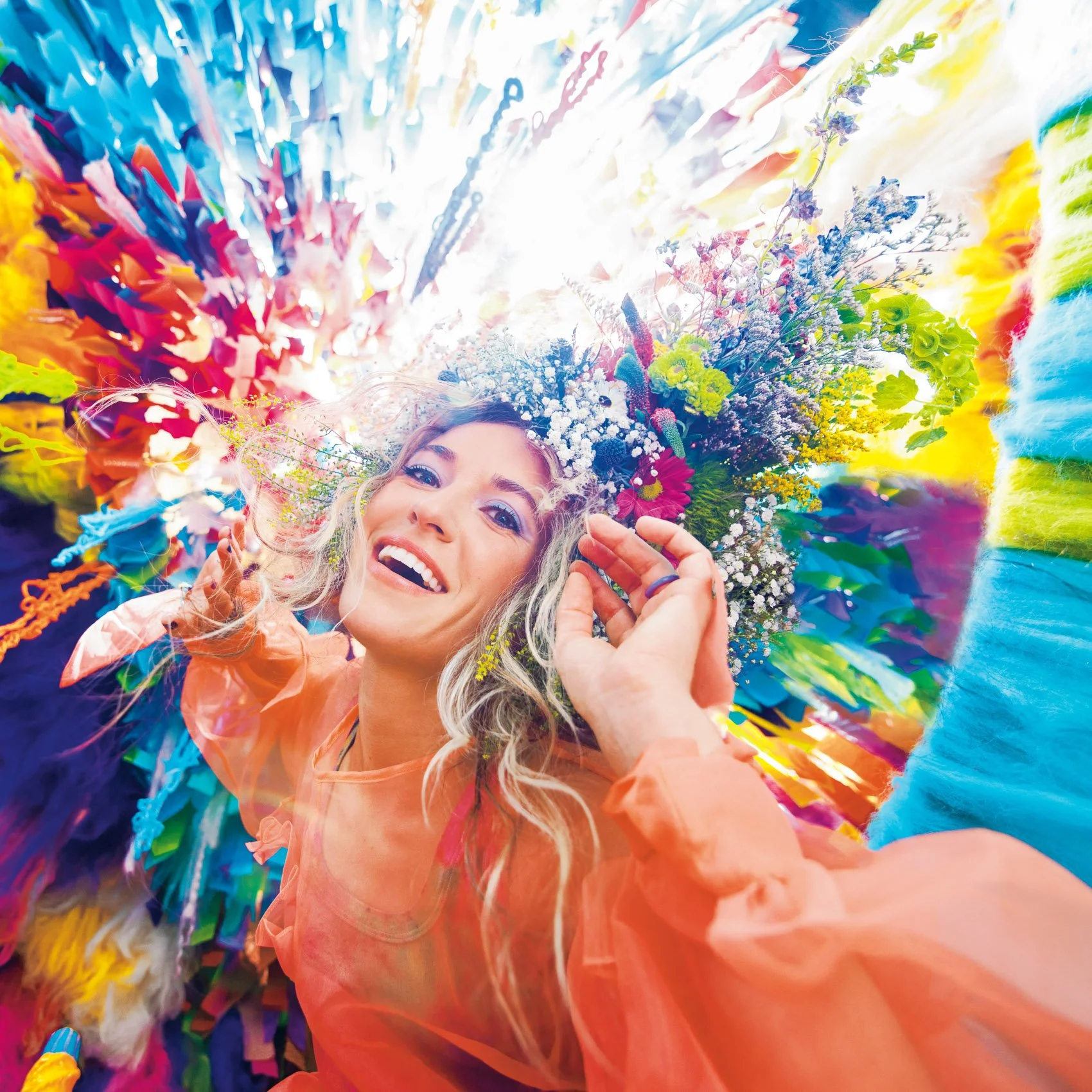 A young woman with blonde hair smiling and lying among colorful flowers and fabric, looking at the camera.
