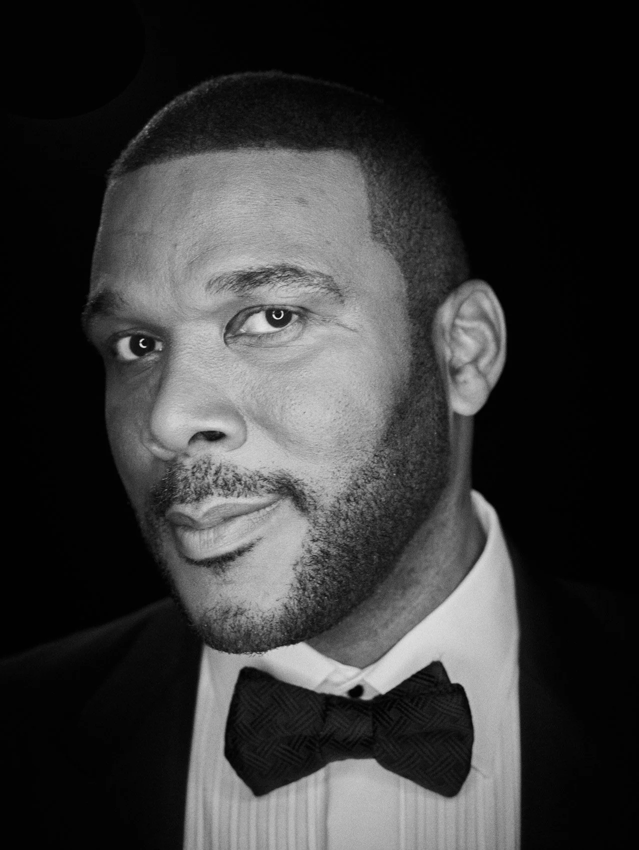 Black and white portrait of a man in formal attire, wearing a tuxedo and bow tie, with short hair and facial hair, looking directly at the camera.
