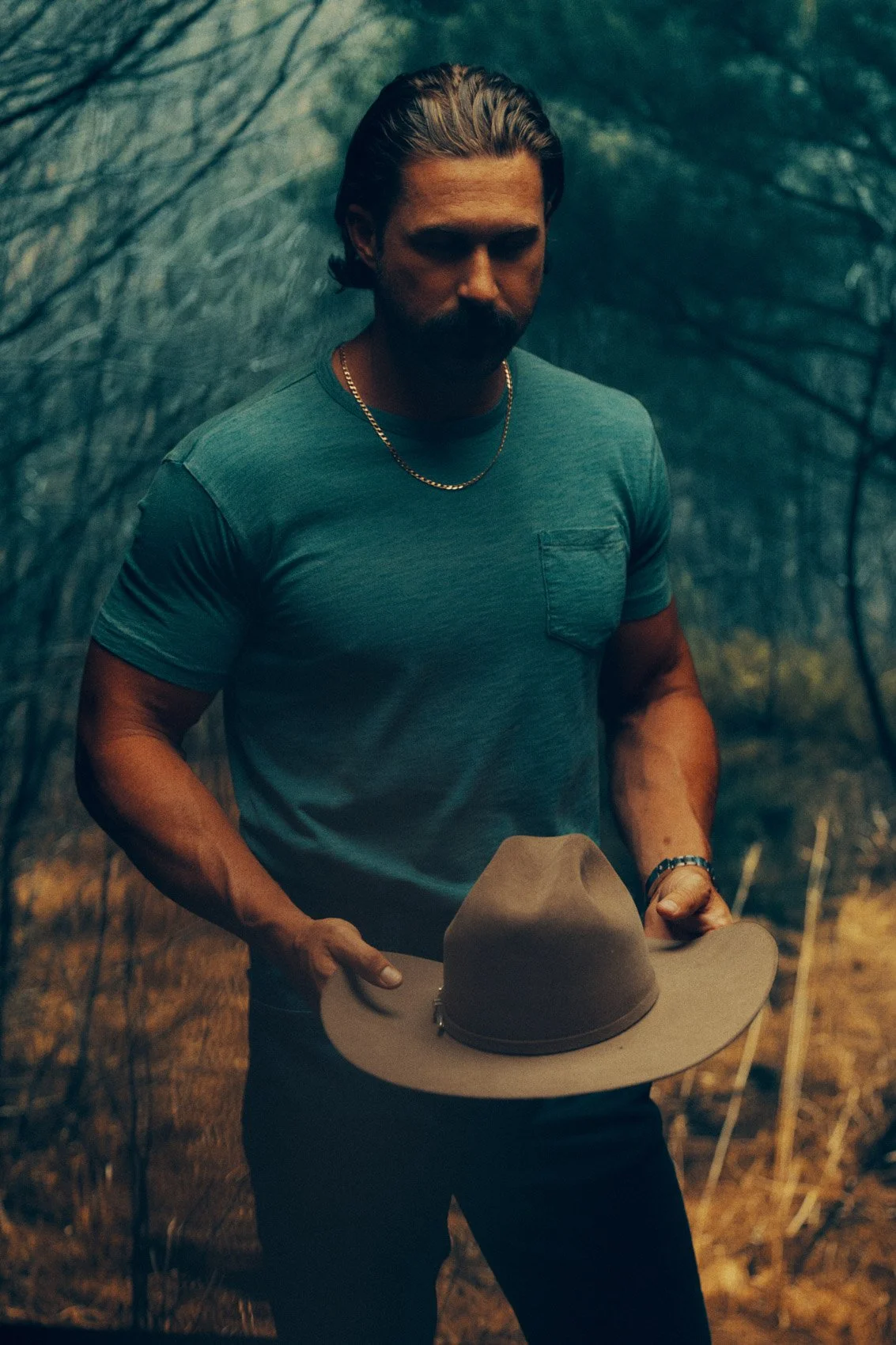 A man in a blue T-shirt holding a beige hat in a wooded outdoor setting.