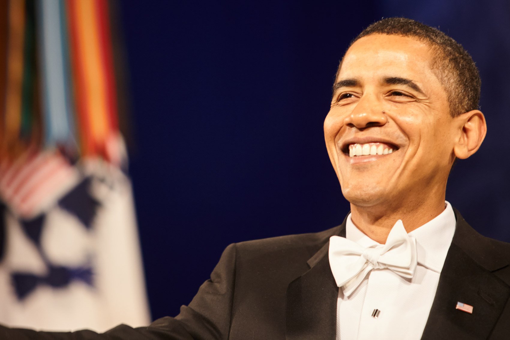 A man in a tuxedo smiling during a formal event, with the American flag in the background.